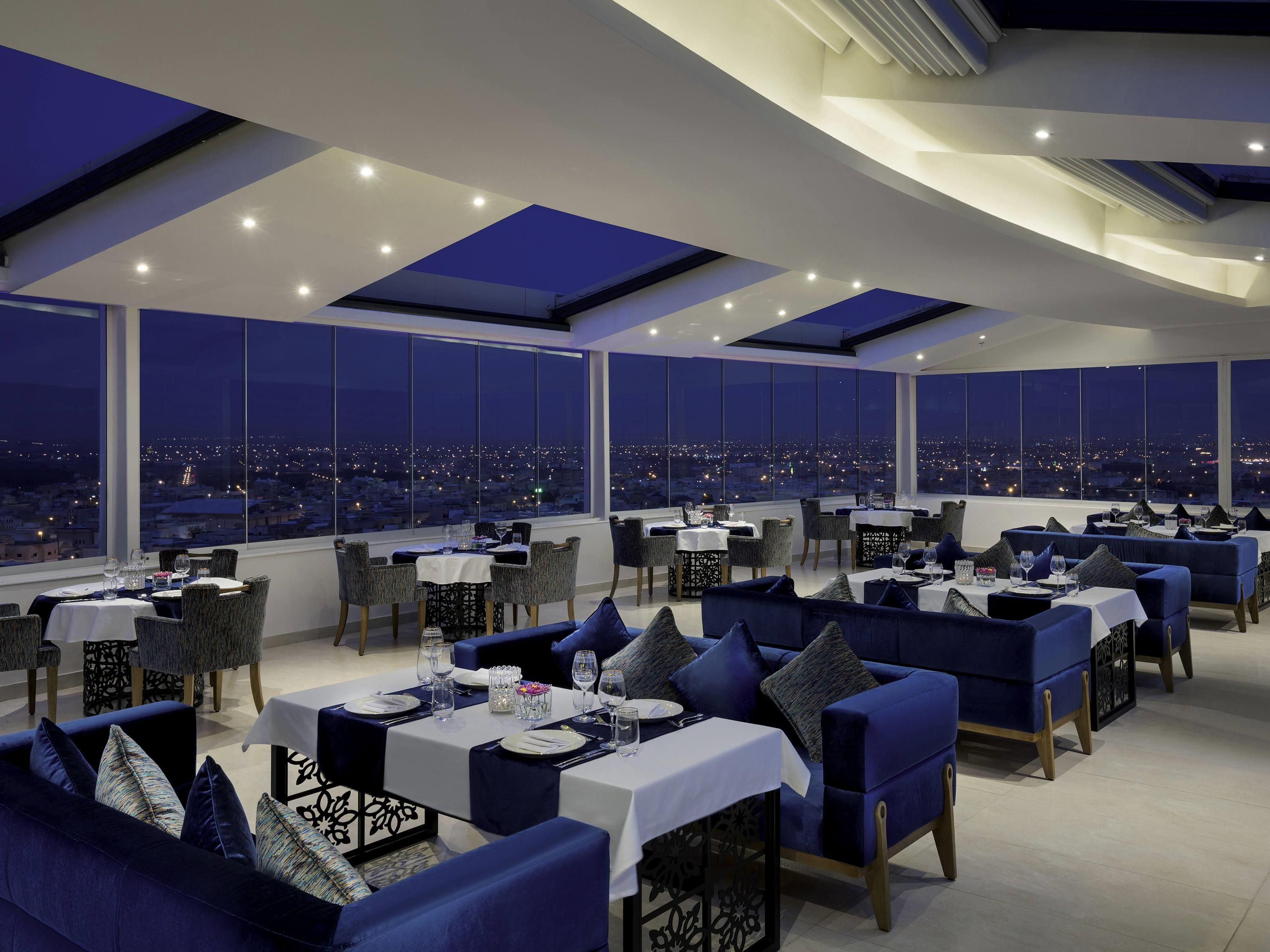 A rooftop restaurant with tables and chairs, large windows offering a city view, and a modern ceiling design.