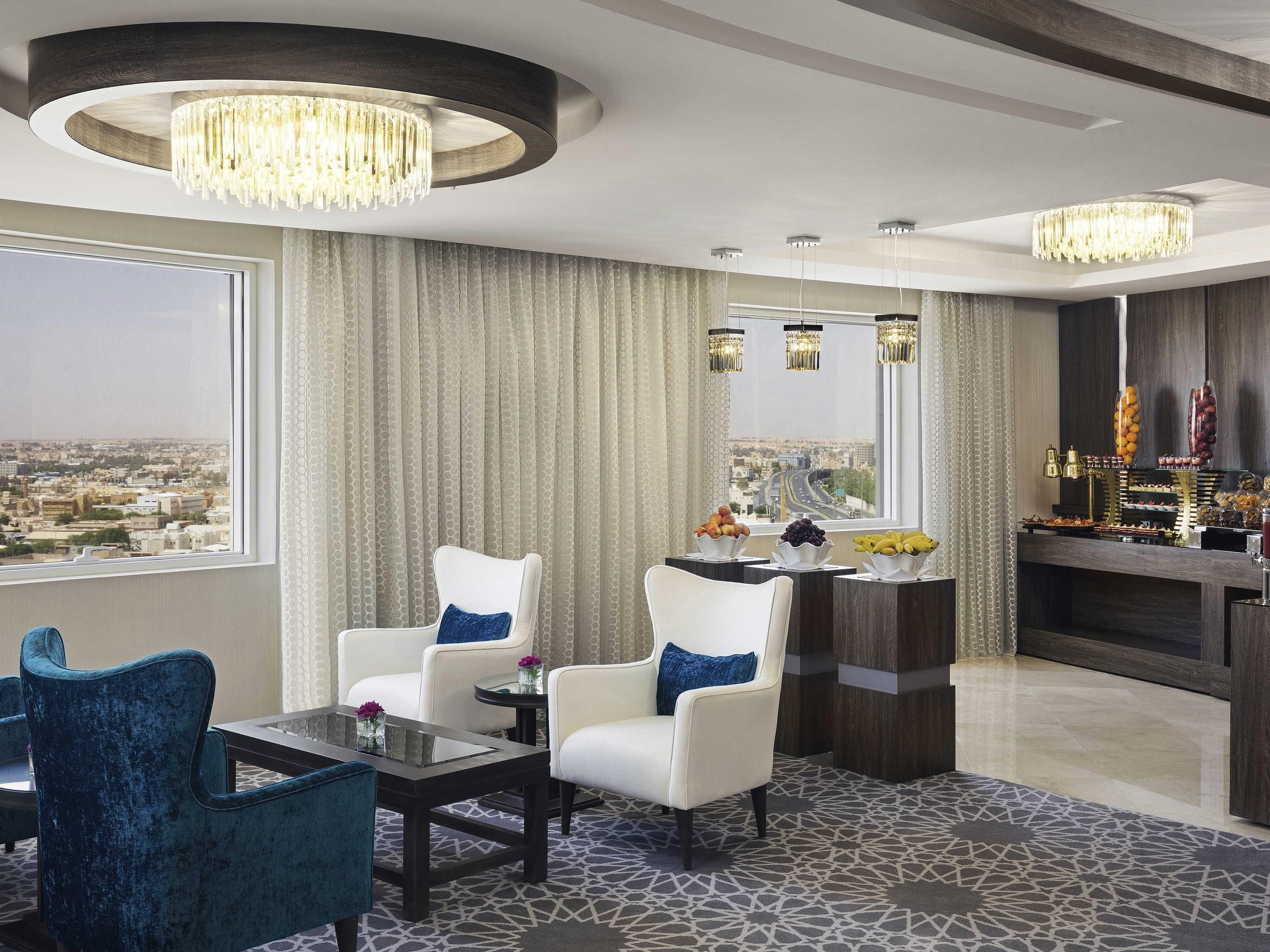 A modern hotel room with a city view, a round chandelier, a bar with fruit and snacks, and a patterned carpet.