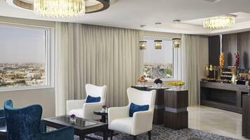 A modern hotel room with a city view, a round chandelier, a bar with fruit and snacks, and a patterned carpet.