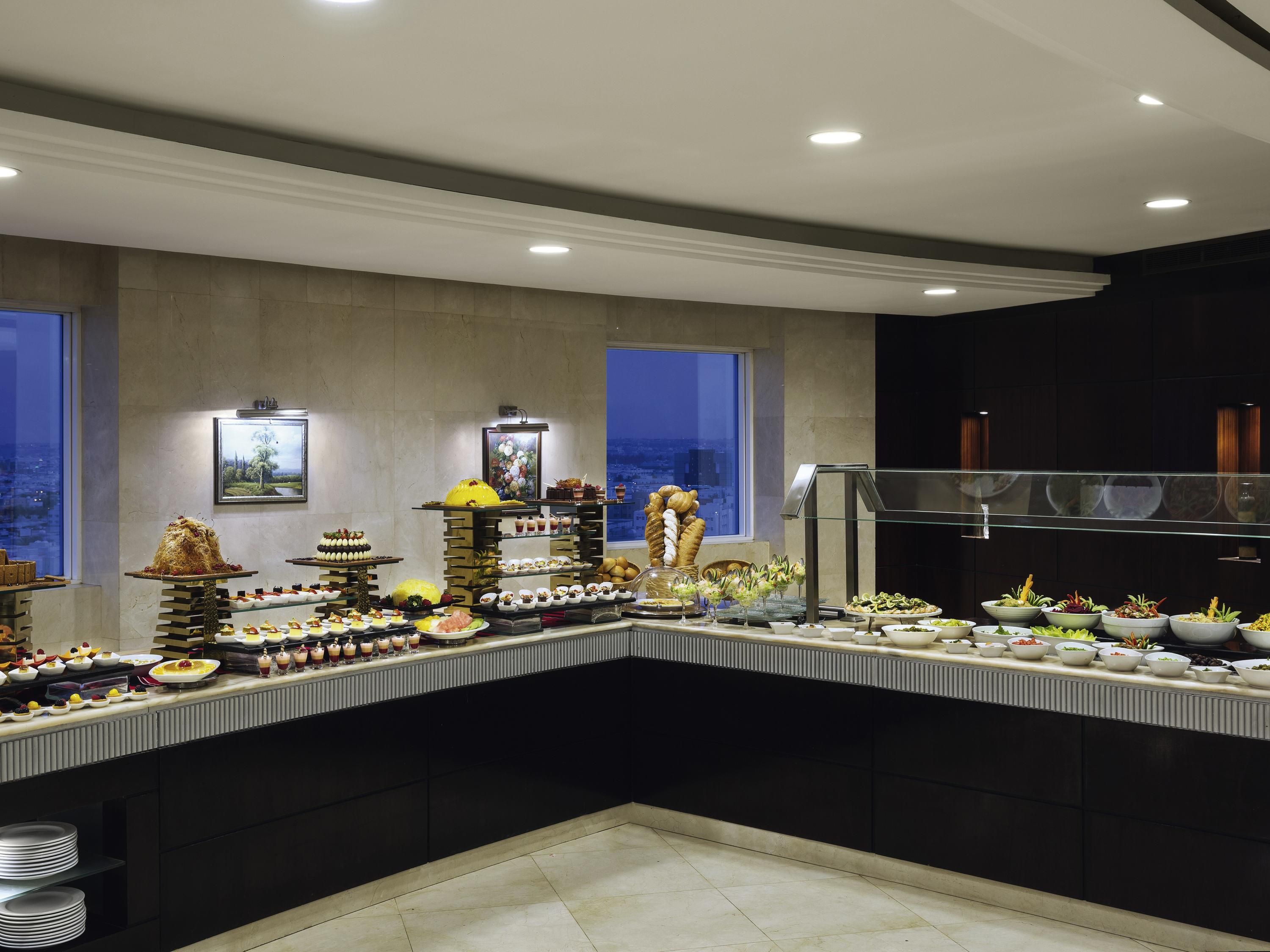 Disposição de buffet com diversos alimentos e bebidas, uma janela ampla com vista para a cidade e design de interiores moderno.