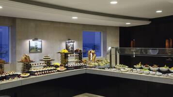 Disposição de buffet com diversos alimentos e bebidas, uma janela ampla com vista para a cidade e design de interiores moderno.