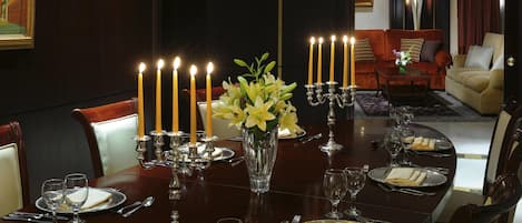 A formal dining room with a long table set for a meal, featuring a candelabra with lit candles, a vase of flowers, and a painting on the wall.