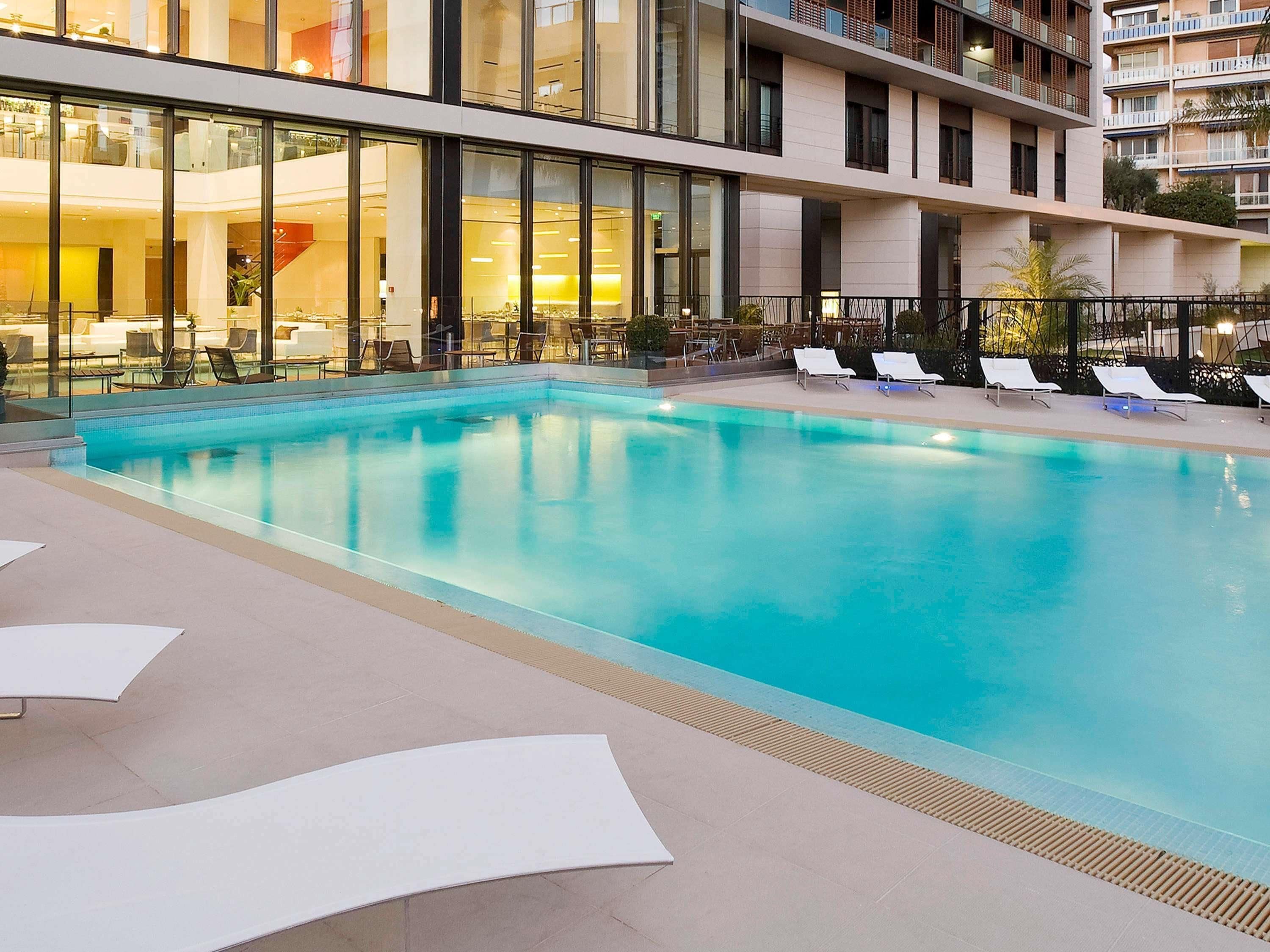 A modern indoor swimming pool with clear blue water, surrounded by lounge chairs and a glass wall.