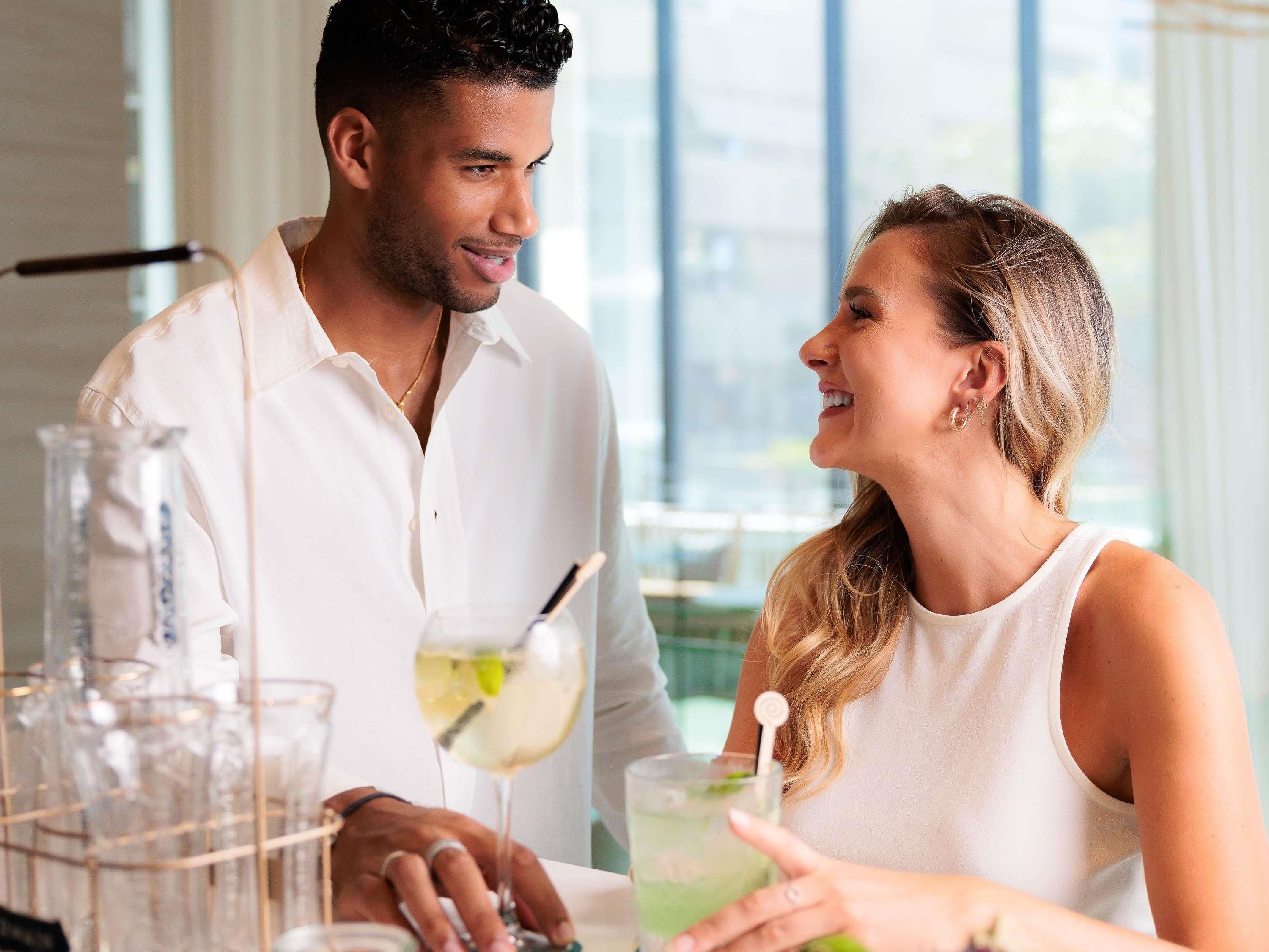 Un homme et une femme sont assis à une table où se trouvent des boissons, dans une pièce dotée de grandes fenêtres et d’un intérieur moderne.