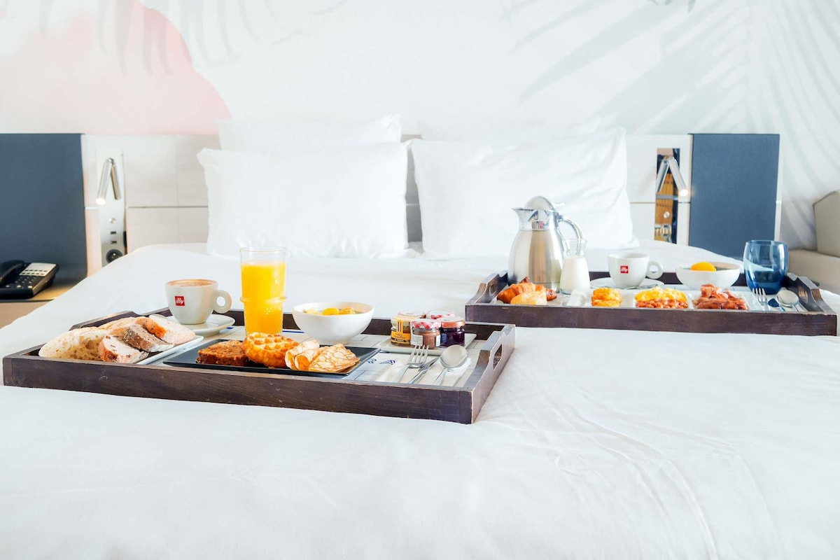 A hotel room with a breakfast tray on the bed, including pastries, a coffee cup, and a glass of orange juice.