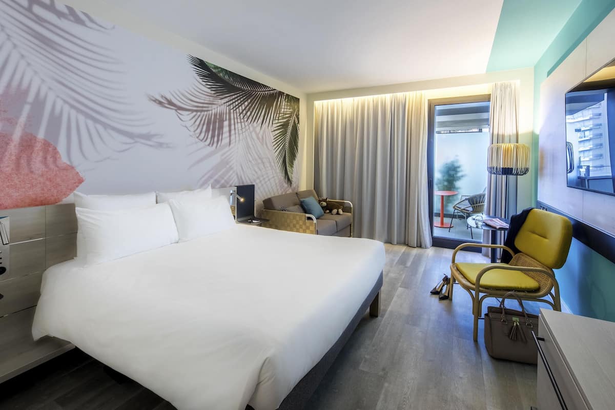 Ein Hotelzimmer mit einem großen Bett, einer Couch, einem Sessel, einem Schreibtisch und Blick ins Freie.