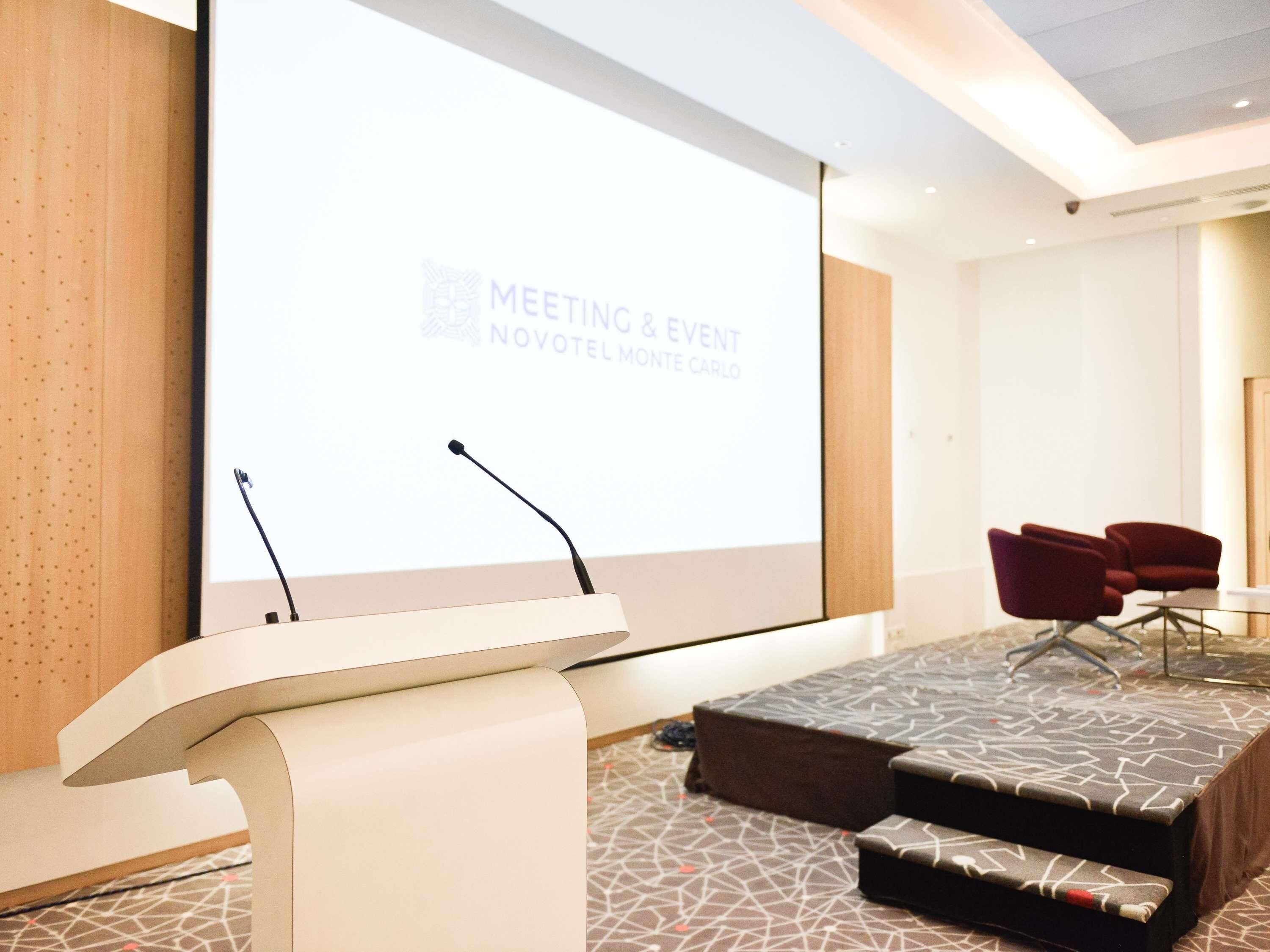 Een vergaderzaal met een spreekgestoelte, een groot scherm waarop 'MEETING & EVENT NOVOTEL MONTE CARLO' staat, en een zithoek met stoelen.