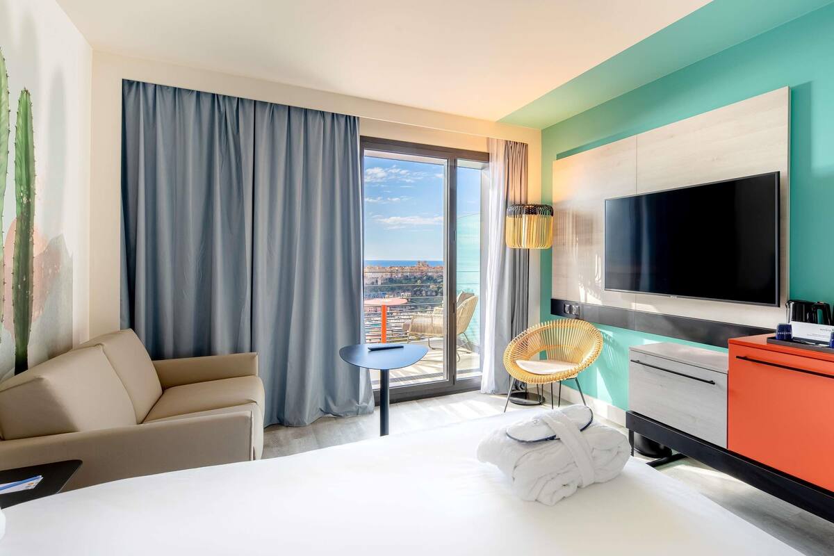 Ein Hotelzimmer mit Bett, Fernseher, Schreibtisch, Sessel und Blick auf die Stadt.