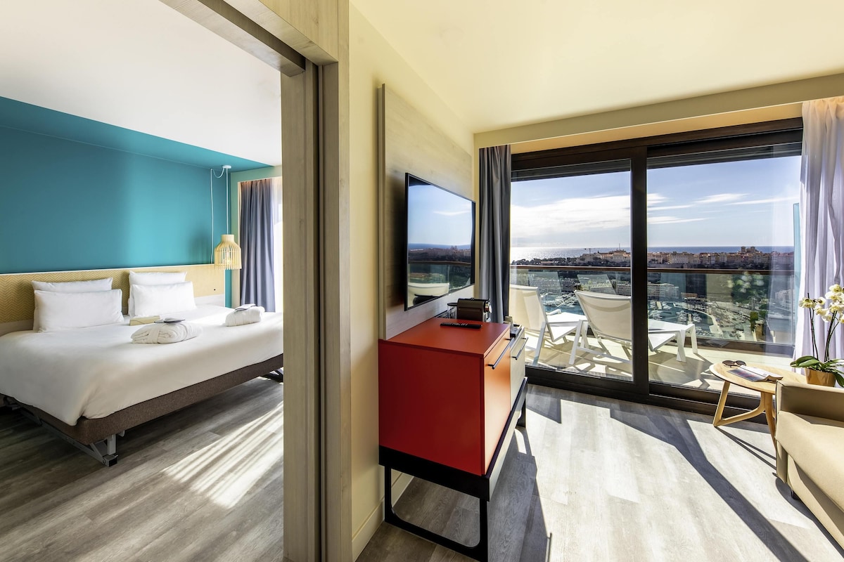 A modern hotel room with a large bed, a desk, and a balcony with a city view.