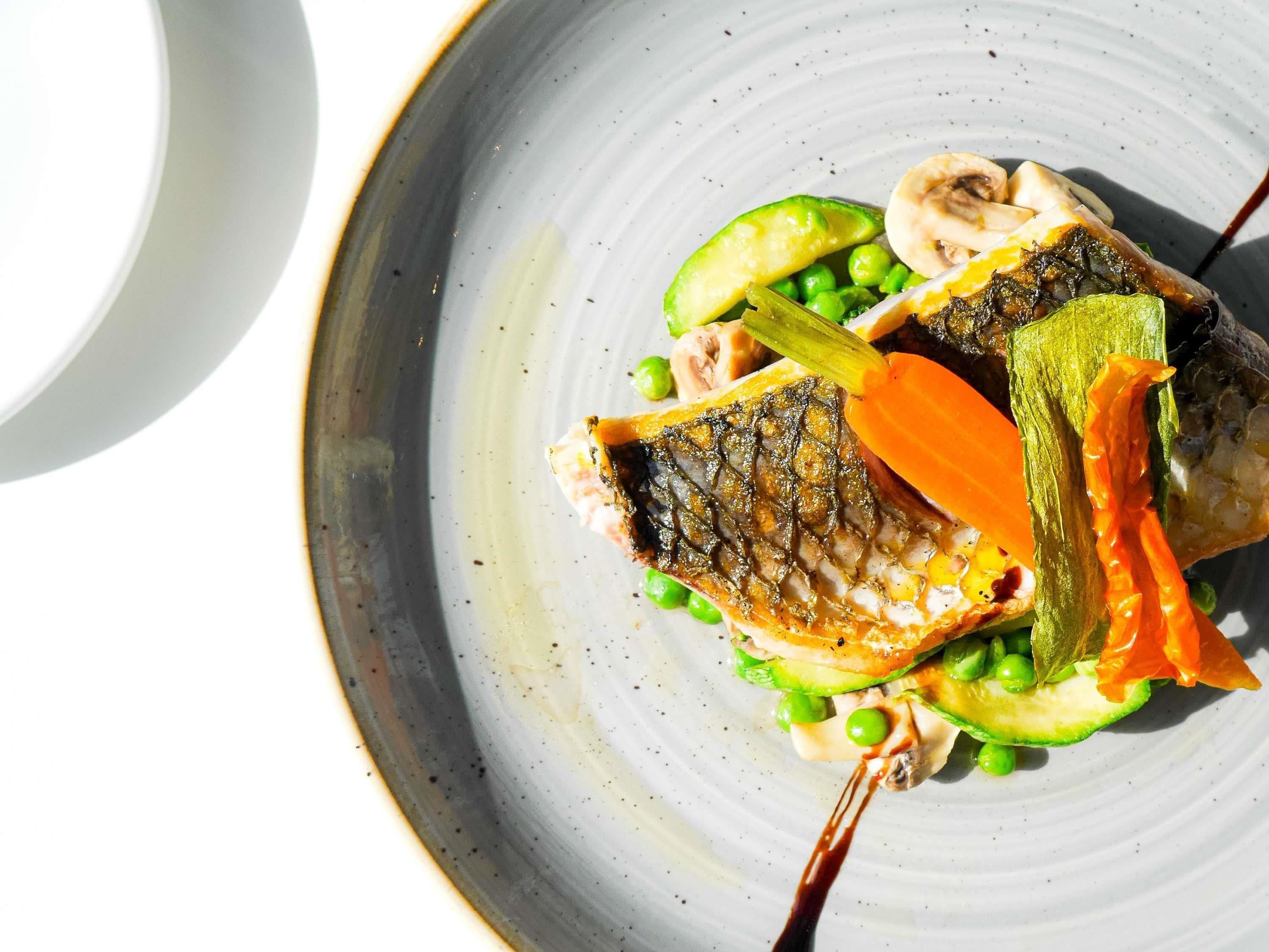A plate with a grilled fish, avocado, peas, and carrot slices.