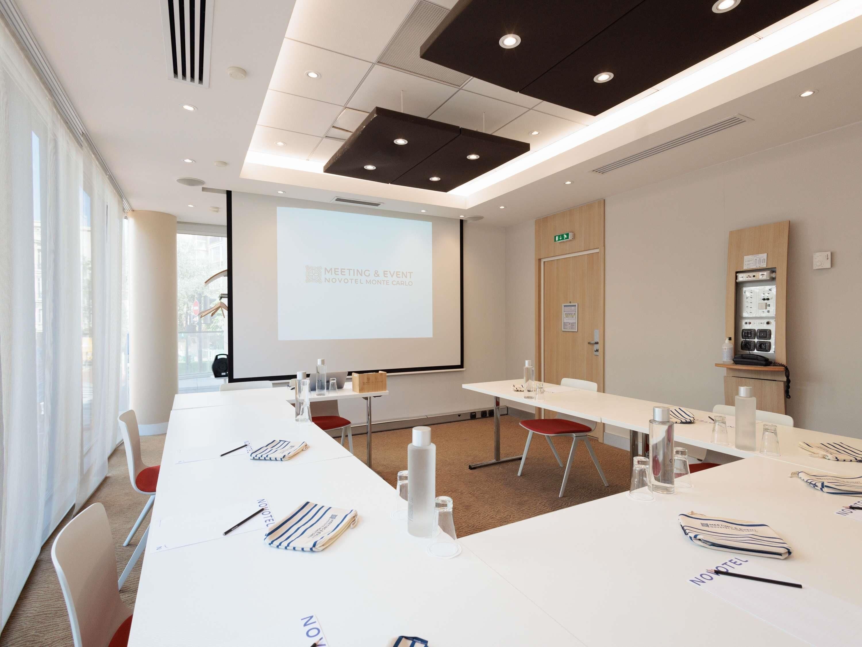 A modern meeting room with white tables, red chairs, and a projector screen displaying 'Meeting & Event Novotel Monte Carlo'.