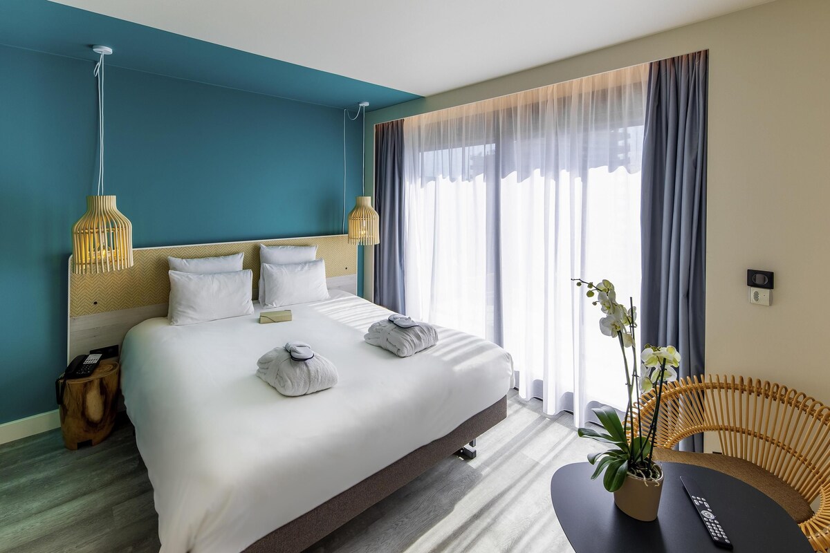 A hotel room with a large bed, bedside lamps, a bedside table, a chair, and a potted plant.