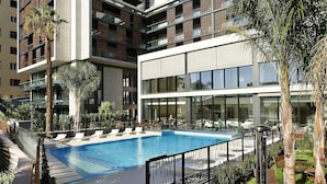 Un hotel moderno de varios pisos con piscina en la azotea, mobiliario exterior y jardines exuberantes.