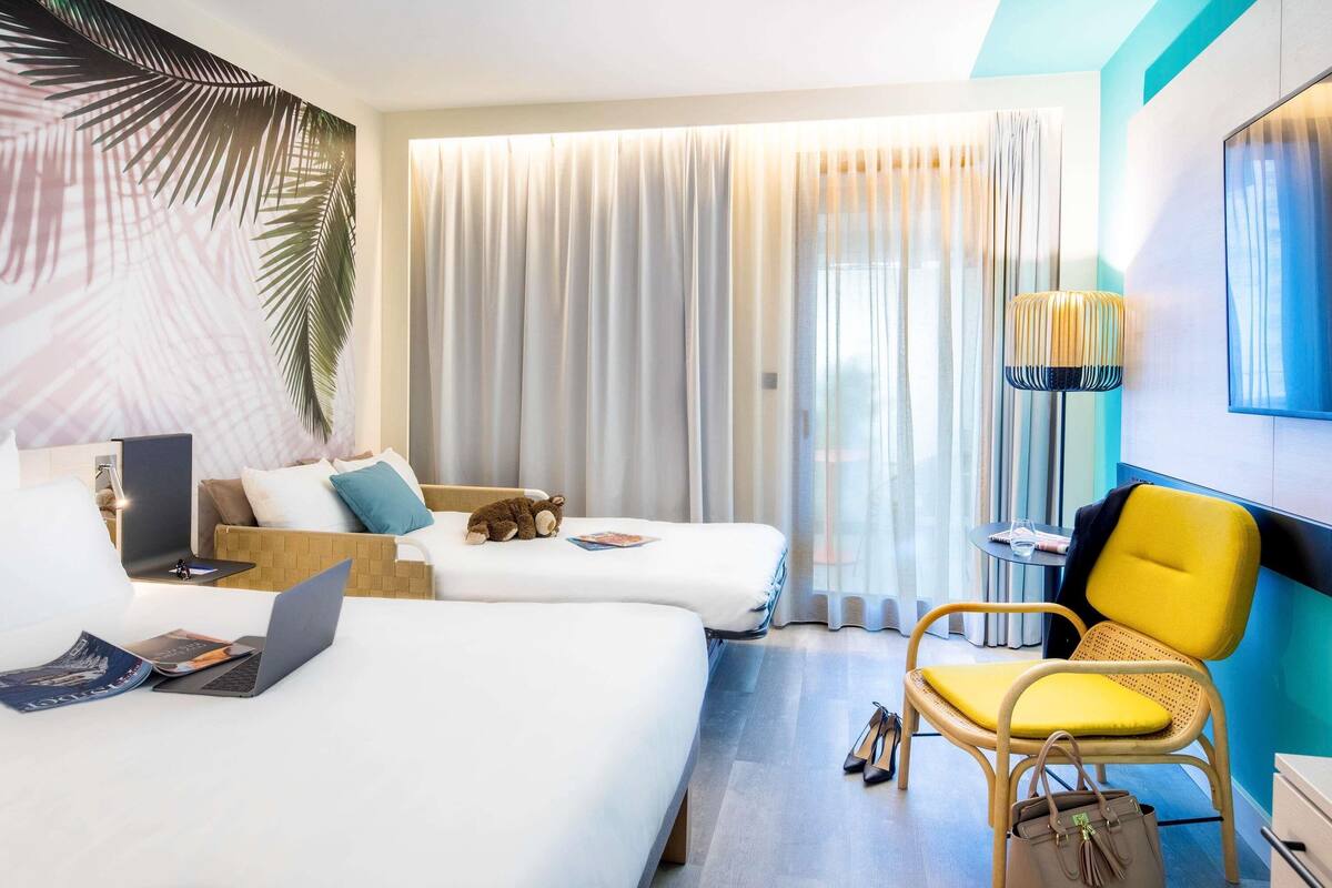 Ein Hotelzimmer mit zwei Betten, einem Schreibtisch, einem Laptop, einem Stuhl und einer Lampe.