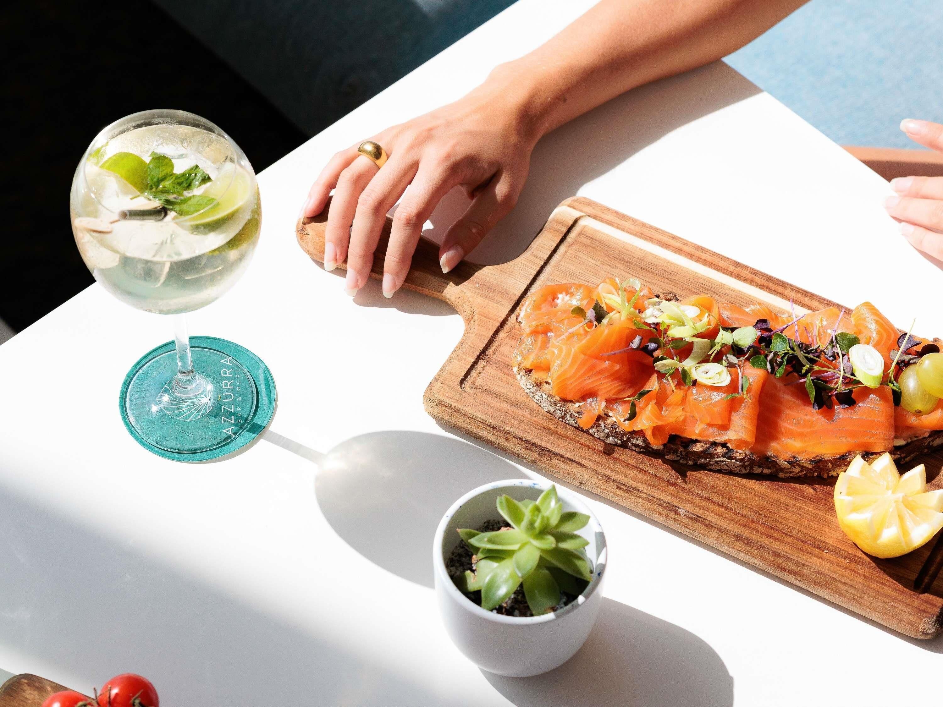 A table with a wooden board holding smoked salmon, a glass of a clear drink with lime, and a small potted plant.