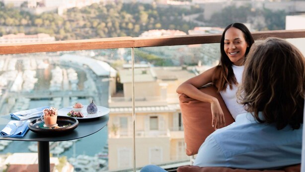 A balcony with a table set for two, overlooking a cityscape with buildings and a bridge.