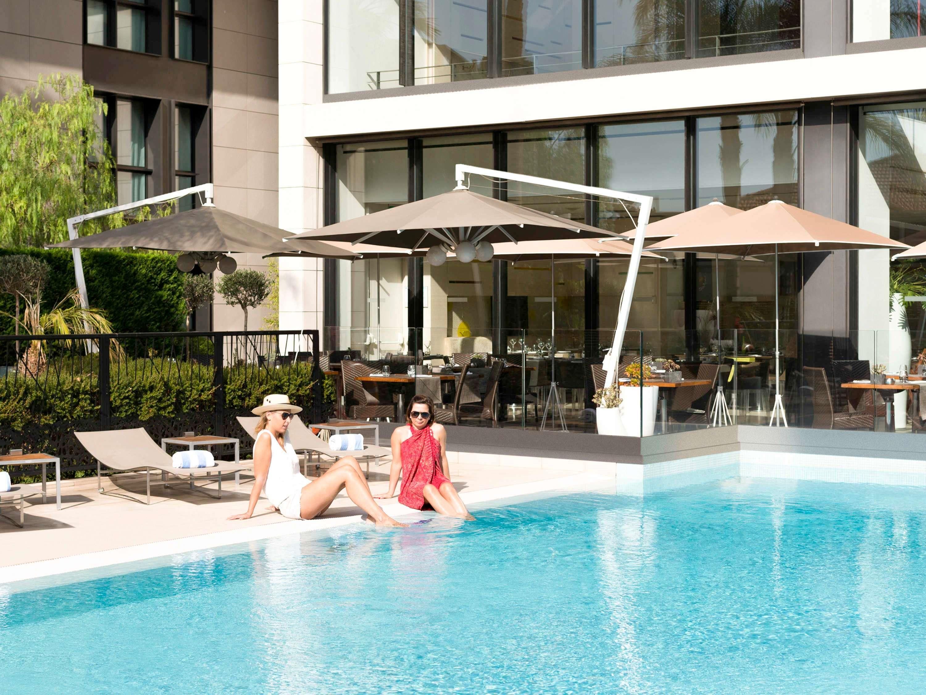 A swimming pool with lounge chairs and umbrellas, surrounded by a modern building with large windows.