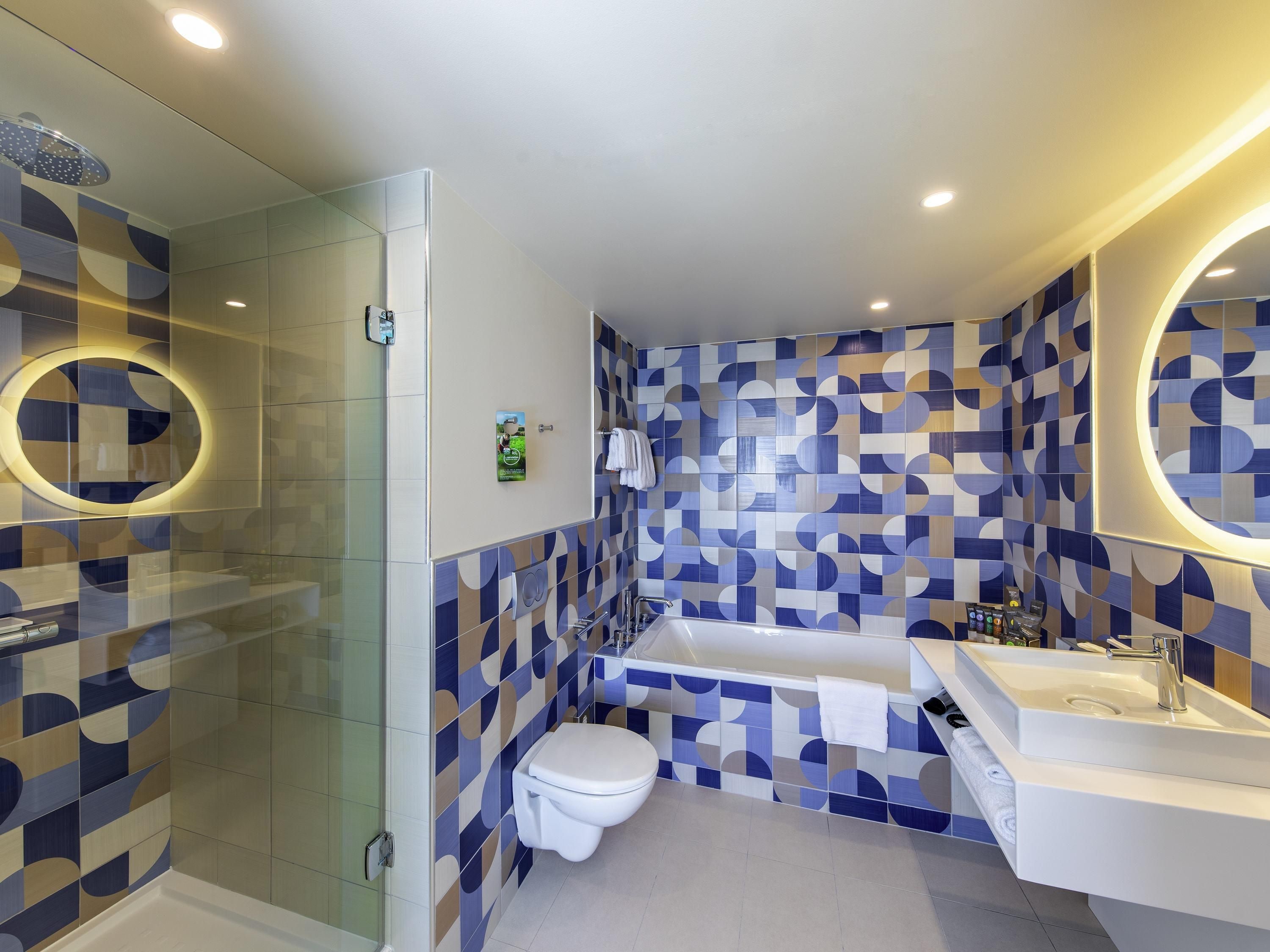Un baño moderno con ducha, inodoro y lavabo, con diseños geométricos en los azulejos y un espejo grande.
