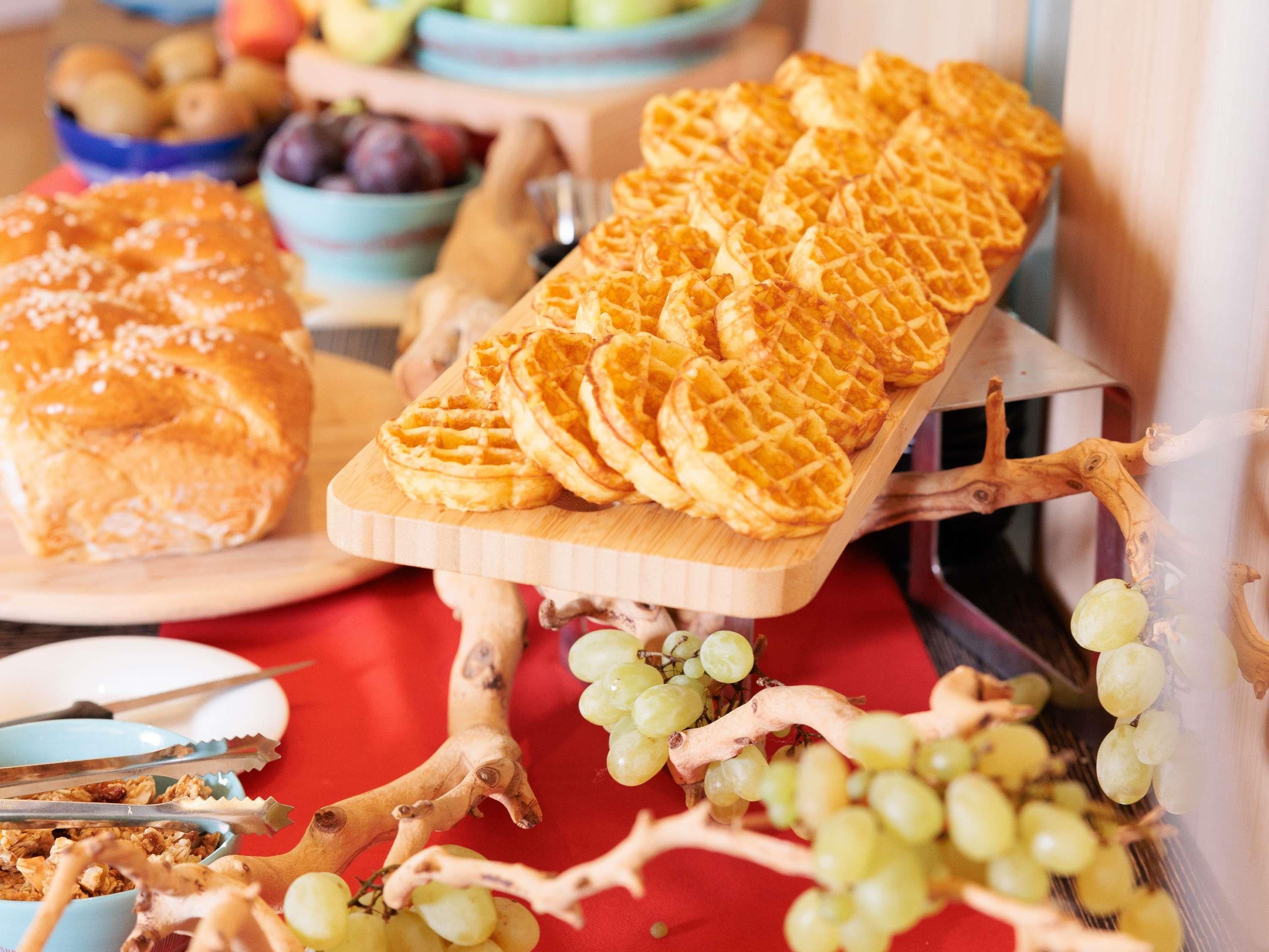 Una tabla de madera con gofres y un pastel, un cuenco de uvas y una frutera al fondo.