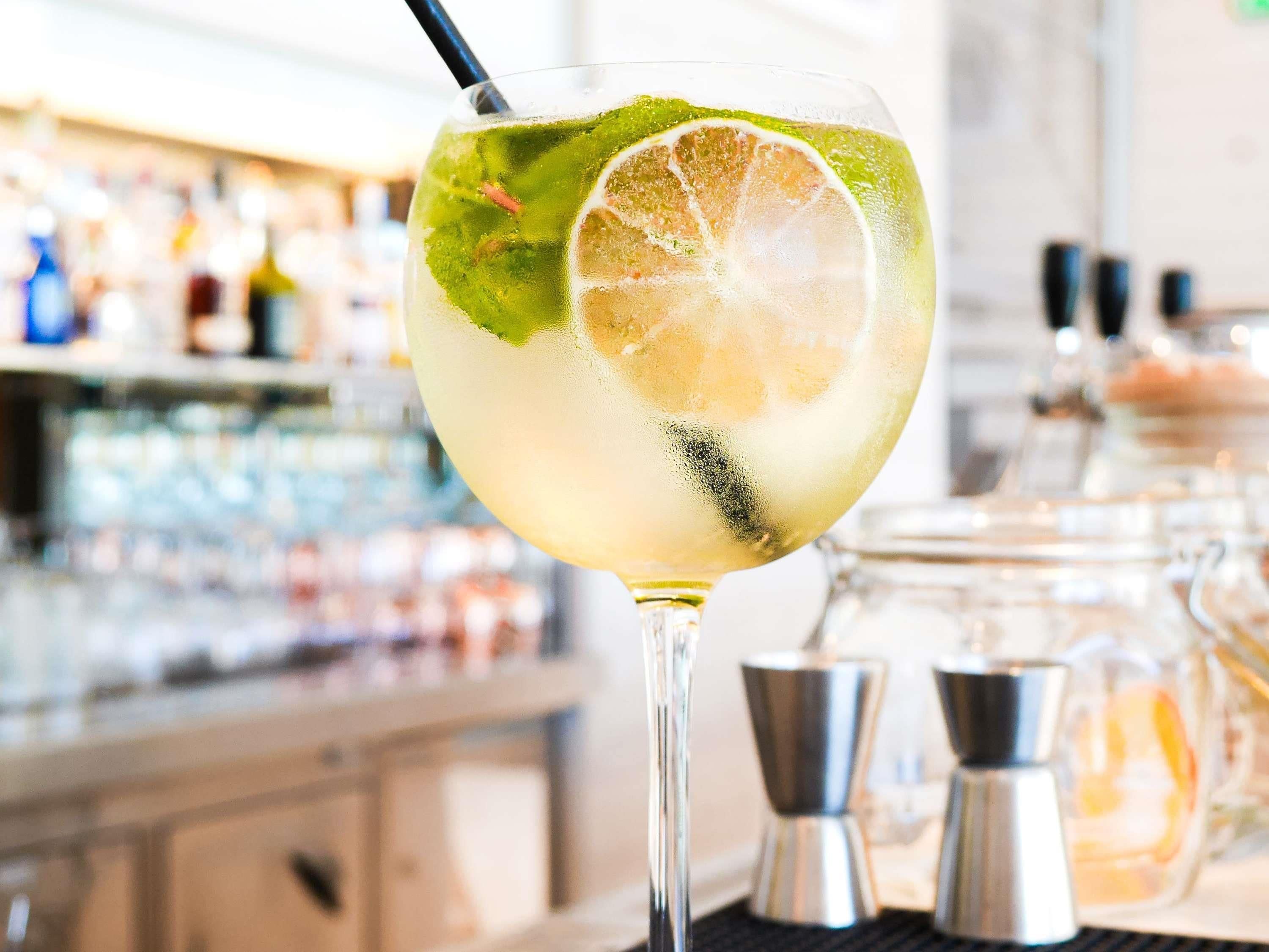 A cocktail glass with a lime slice and mint garnish, placed on a bar counter with a blurred background of shelves and bottles.