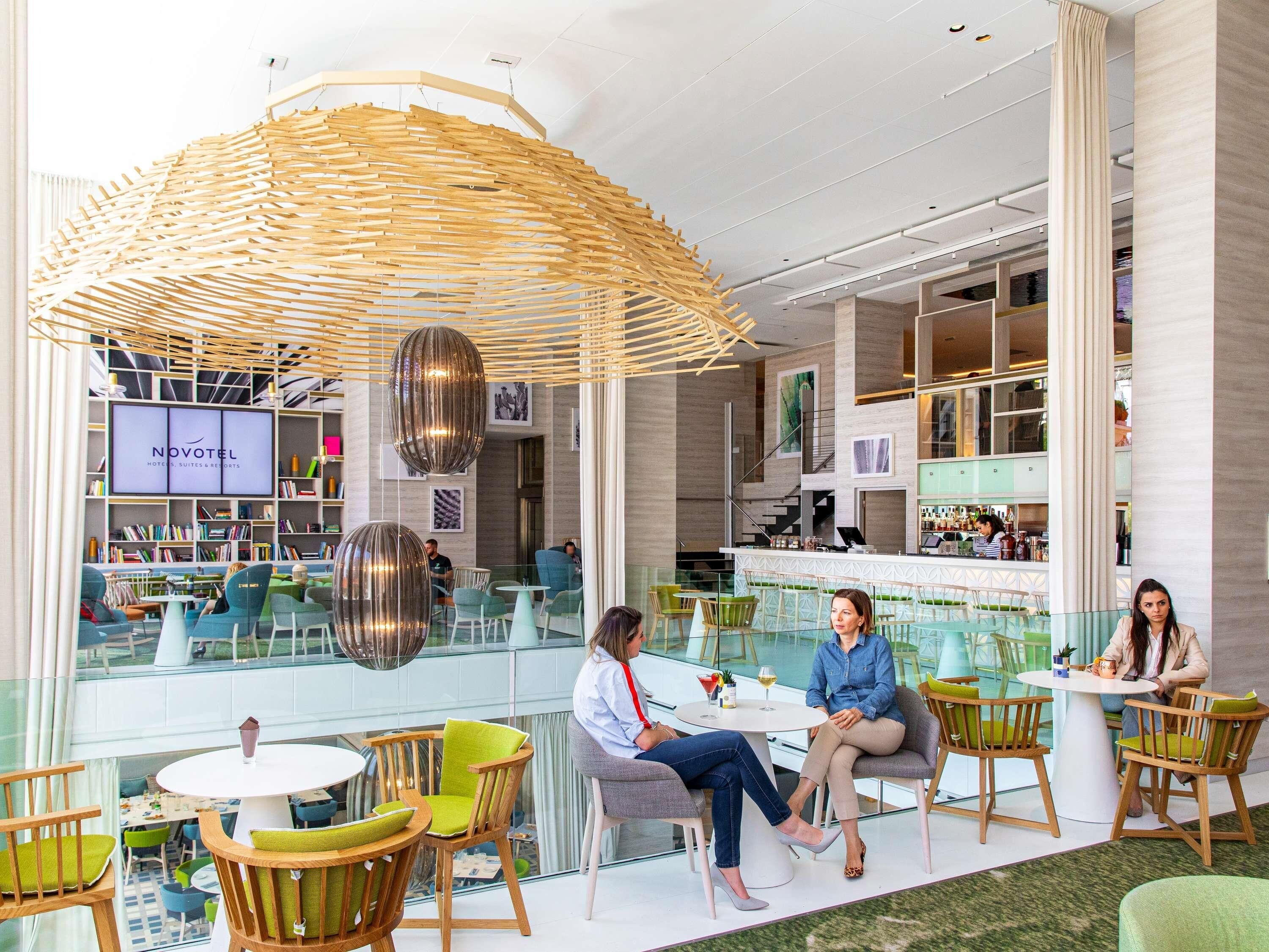 A modern hotel lobby with a central bar, seating areas, and a large screen displaying the hotel name.
