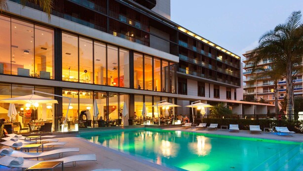 Un hotel con piscina, mobiliario exterior y un edificio de varios pisos con grandes ventanales.