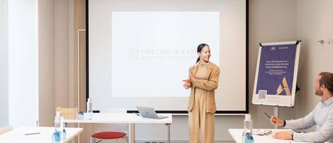 A modern conference room with a presentation screen, a whiteboard, and a table with chairs and laptops.