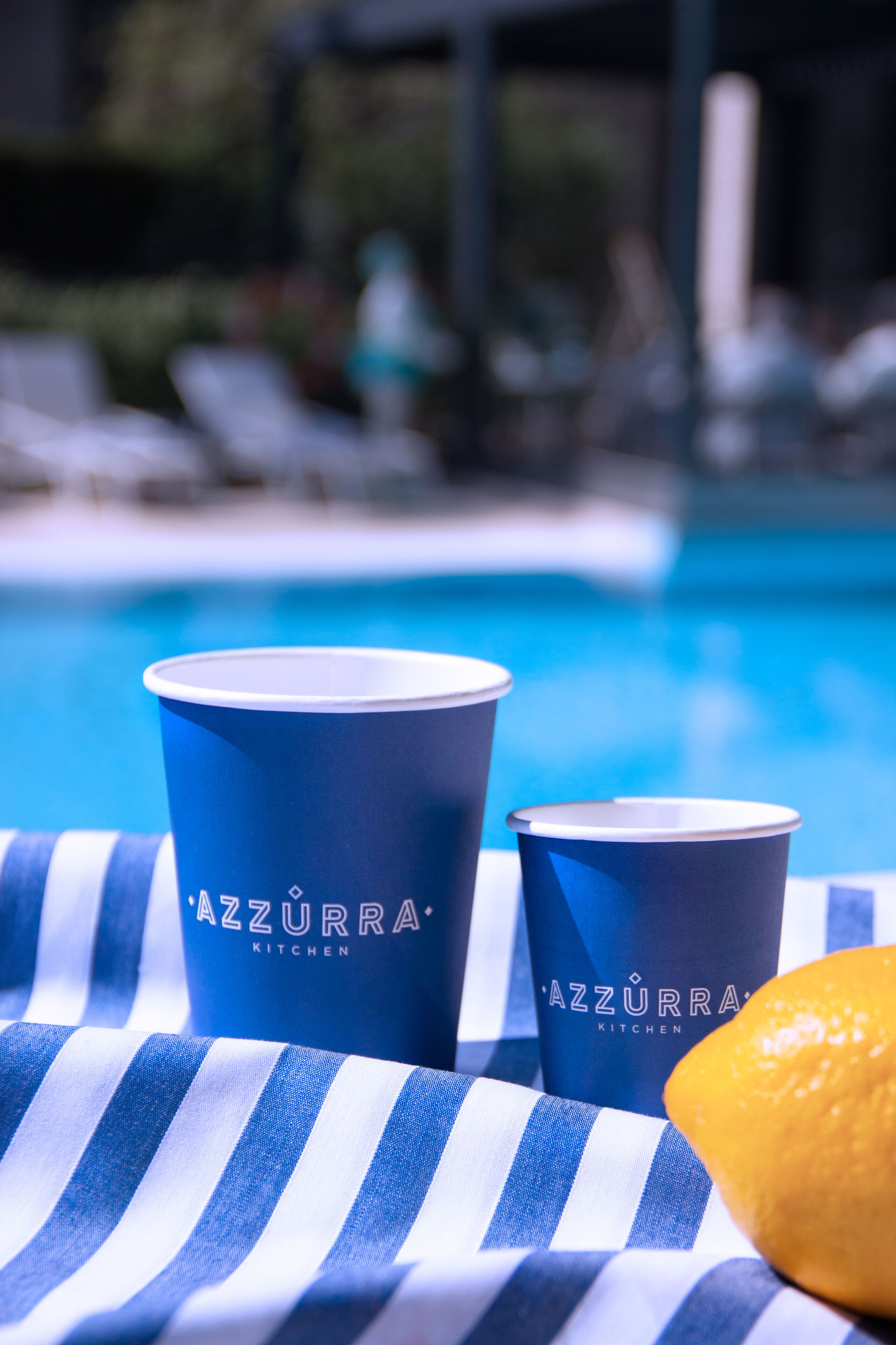 Dos tazas azules con el texto 'Azzurra Kitchen', una toalla azul y blanca a rayas, y un limón en un entorno junto a la piscina.