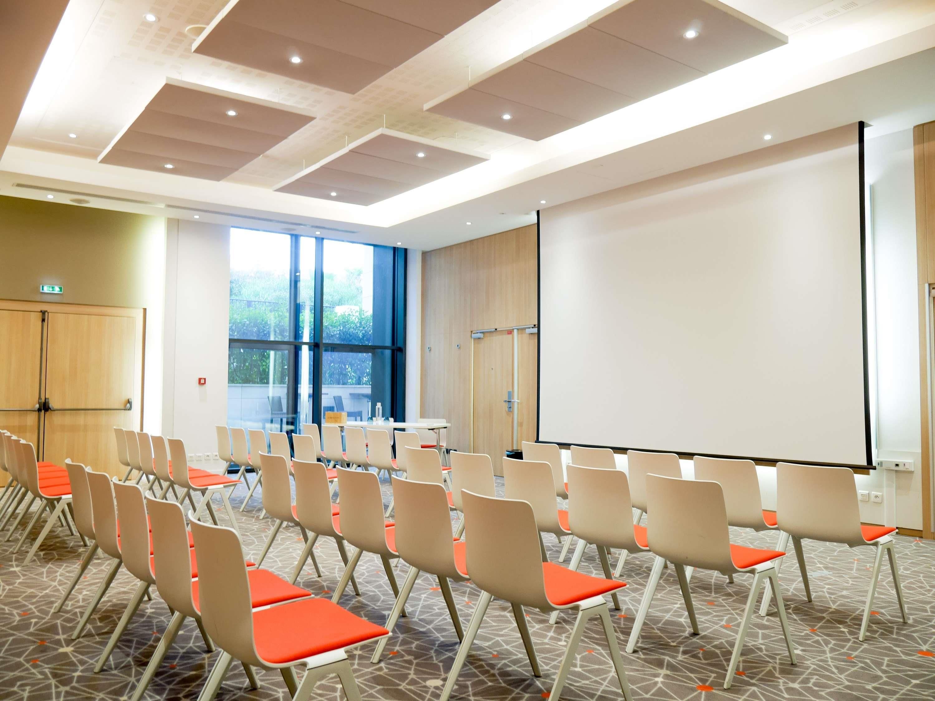 Un salón de conferencias con filas de sillas, una pantalla de proyección y un ventanal grande.