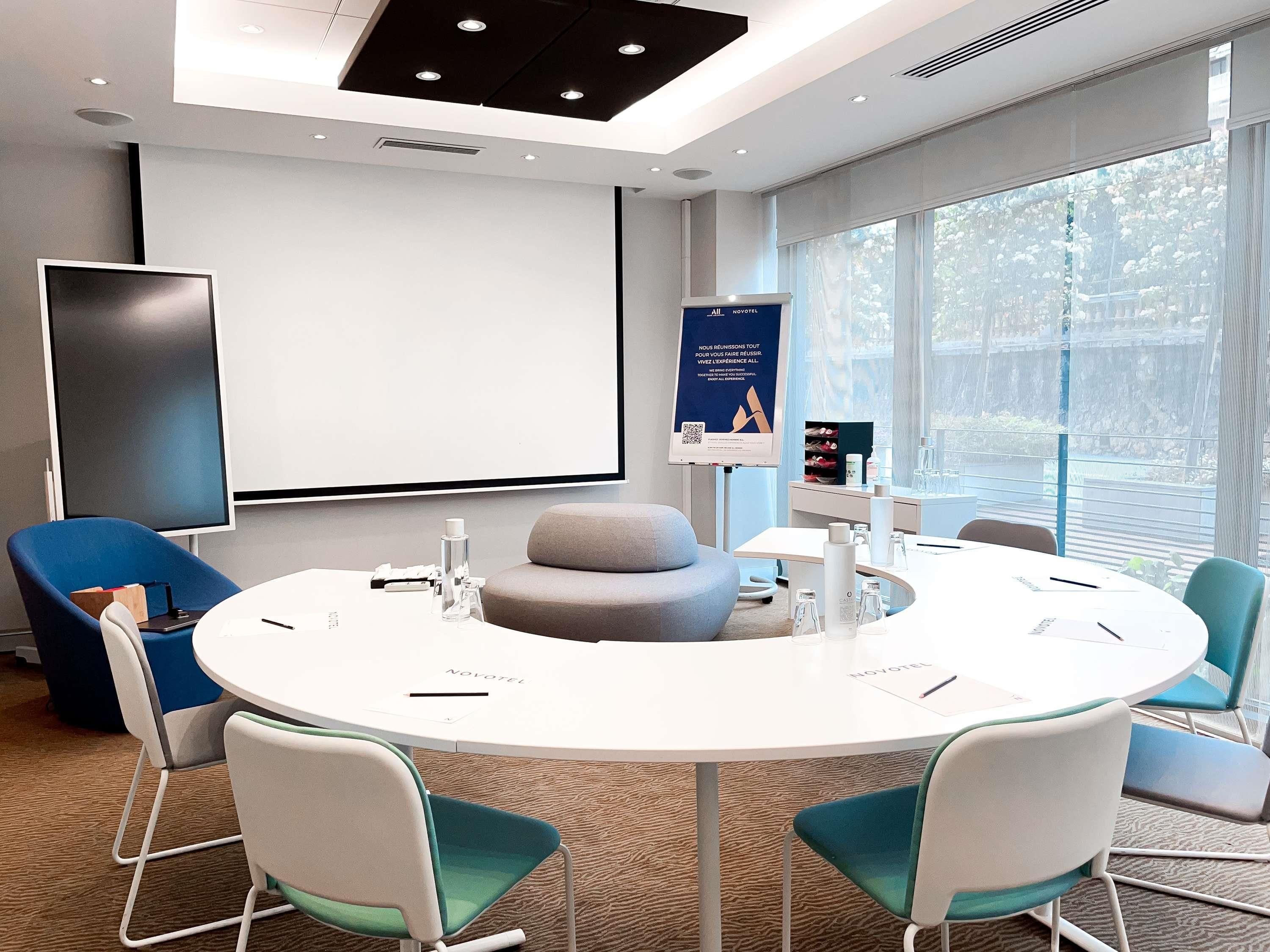 A modern meeting room with a white table, teal chairs, a grey ottoman, and a projector screen.