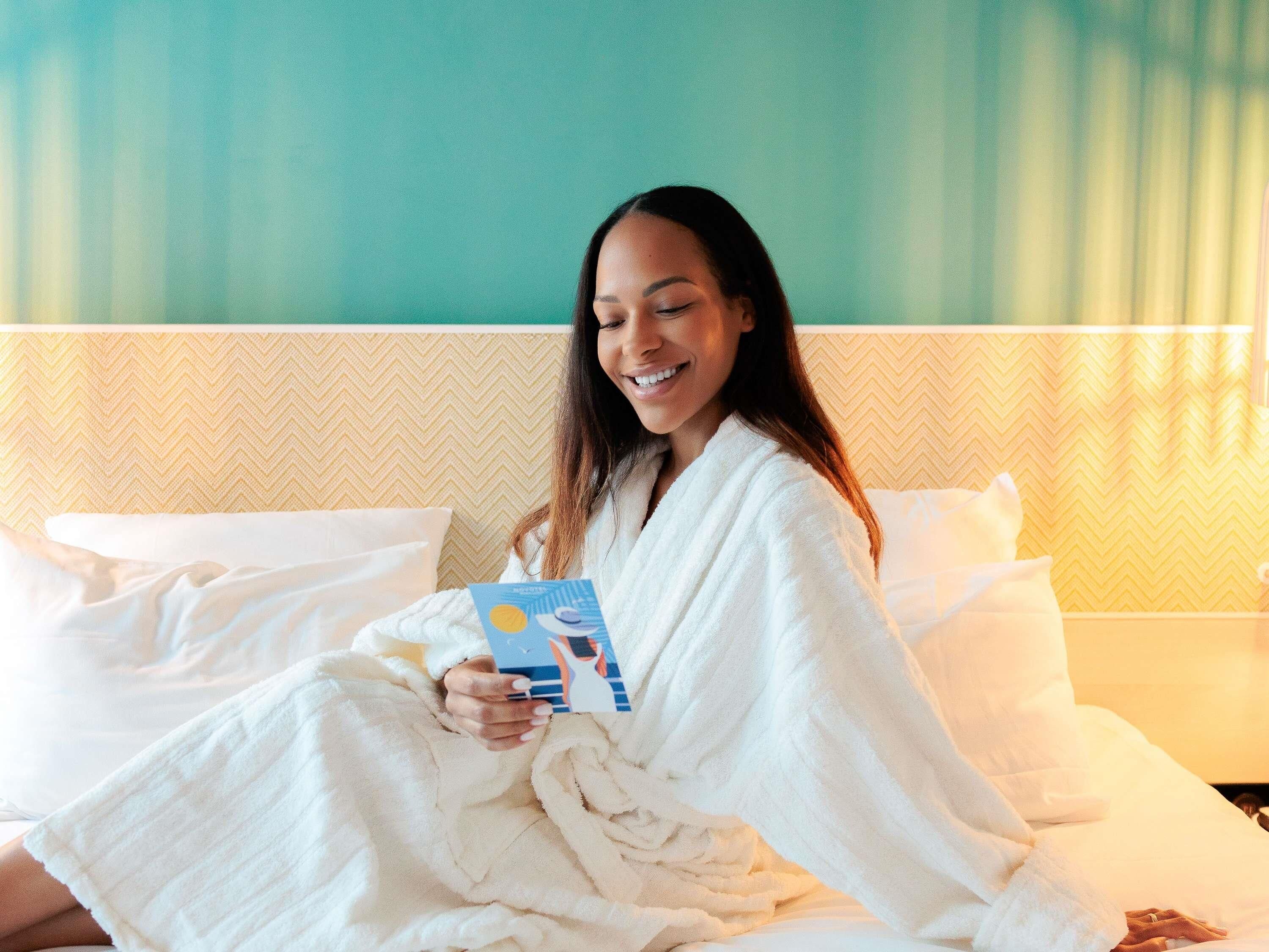 A person in a white bathrobe sitting on a bed with a book, in a room with a patterned wall and a white headboard.