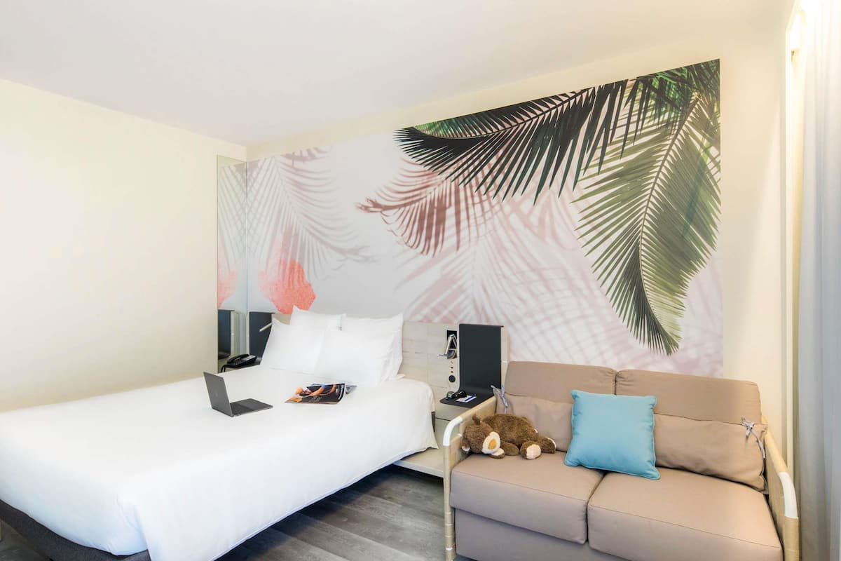 Ein Hotelzimmer mit einem Bett, einem Schlafsofa, einem Nachttisch, einer Lampe und einer Wand mit Blättermotiv.