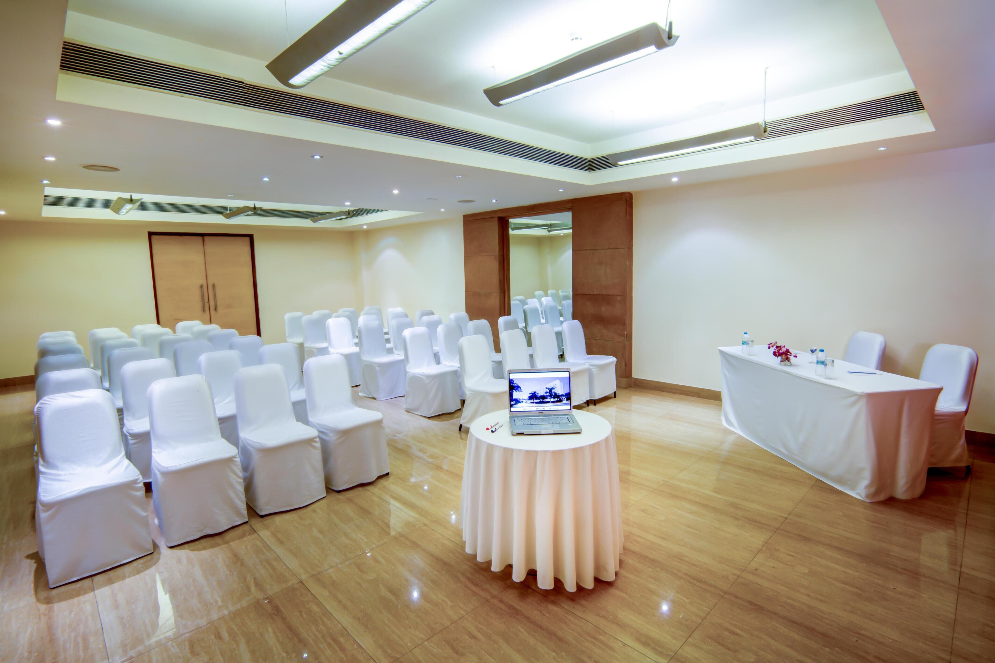 A conference room with rows of white chairs, a round table with a laptop, and a white table with water bottles.
