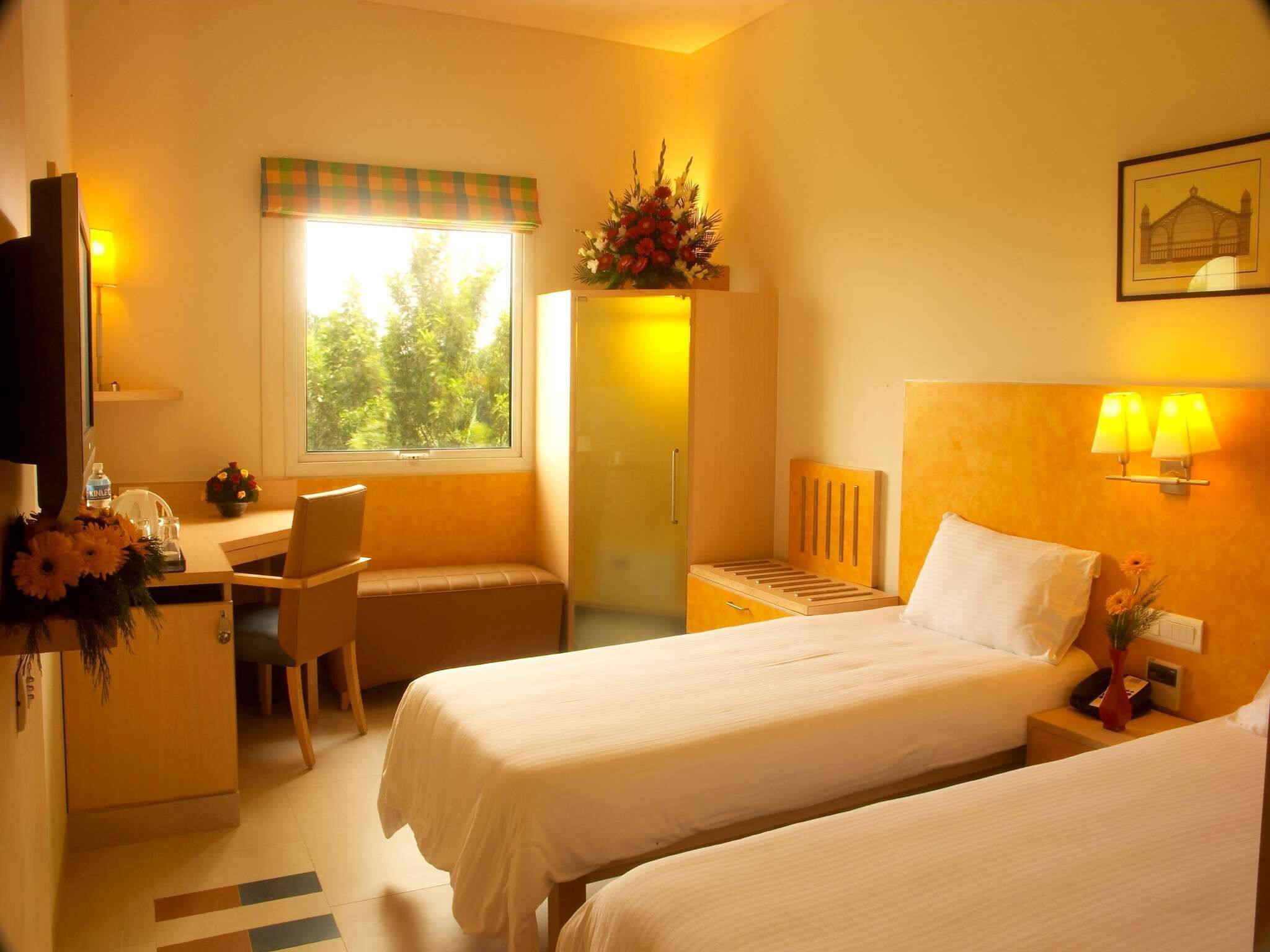 Ein Hotelzimmer mit zwei Betten, einem Schreibtisch, einem Stuhl, einem Fenster mit Blick auf Bäume und einer Wandlampe.
