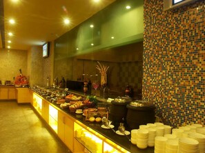 A buffet setup with various food items and utensils, a tiled wall, and a television screen. - Radha Hometel Whitefield (Bengaluru)