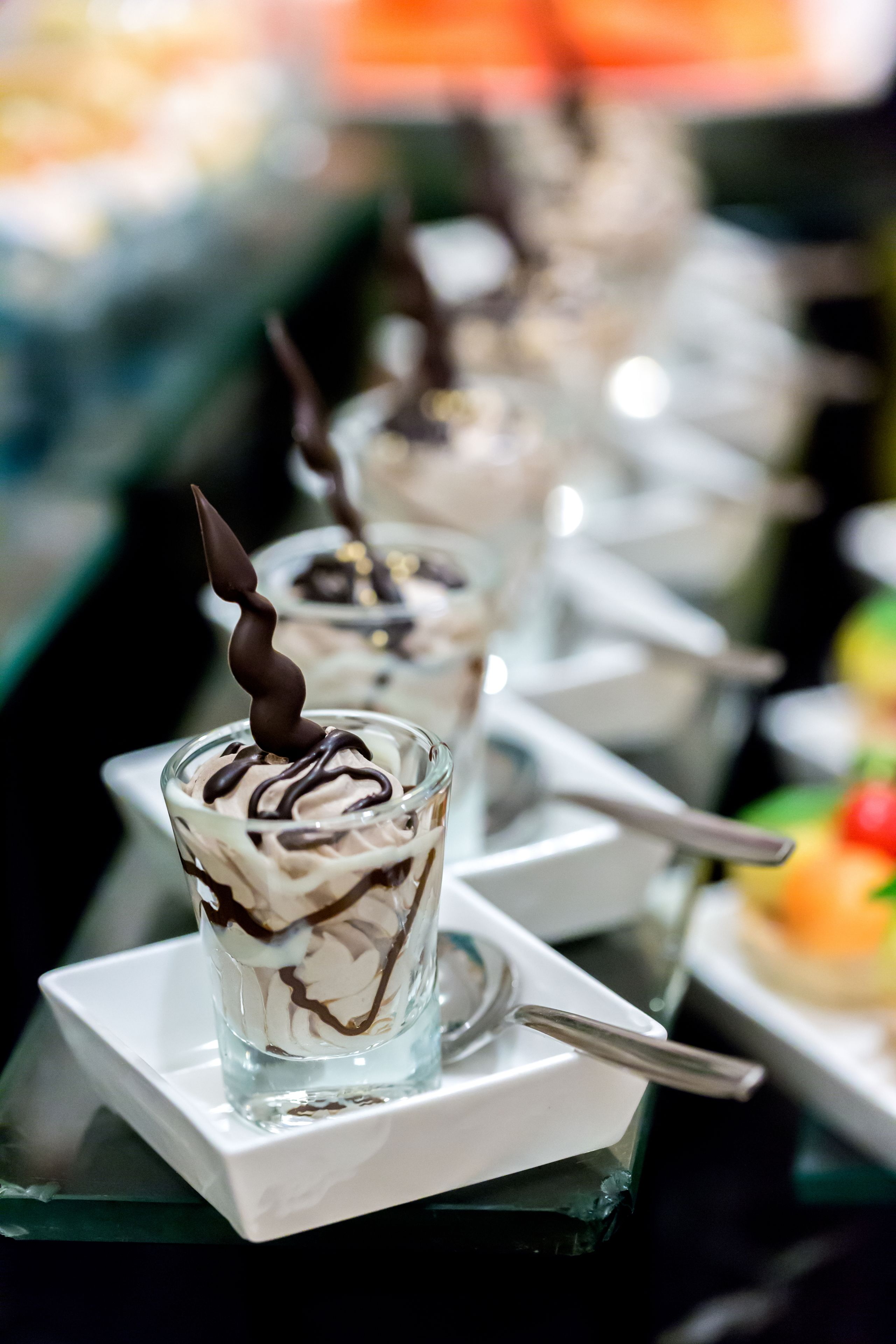 Een dessertpresentatie met chocoladekrullen en een chocoladestukje bovenop, geserveerd in glazen op witte, vierkante borden.