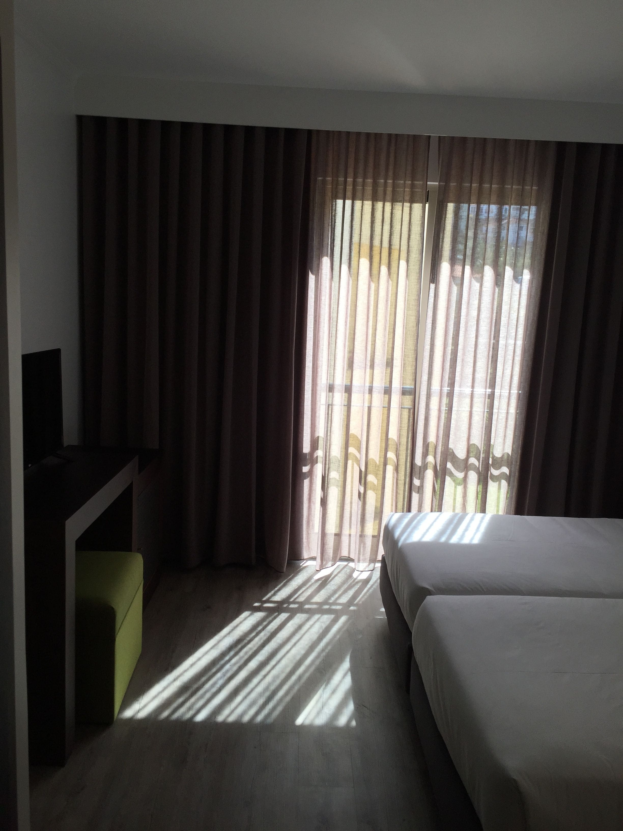 A hotel room with a bed, a desk, and a view of the outside through sheer curtains.