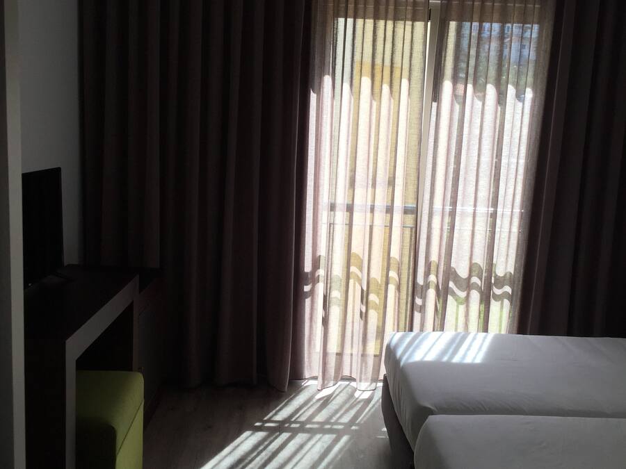 A hotel room with a bed, a desk, and a view of the outside through sheer curtains.