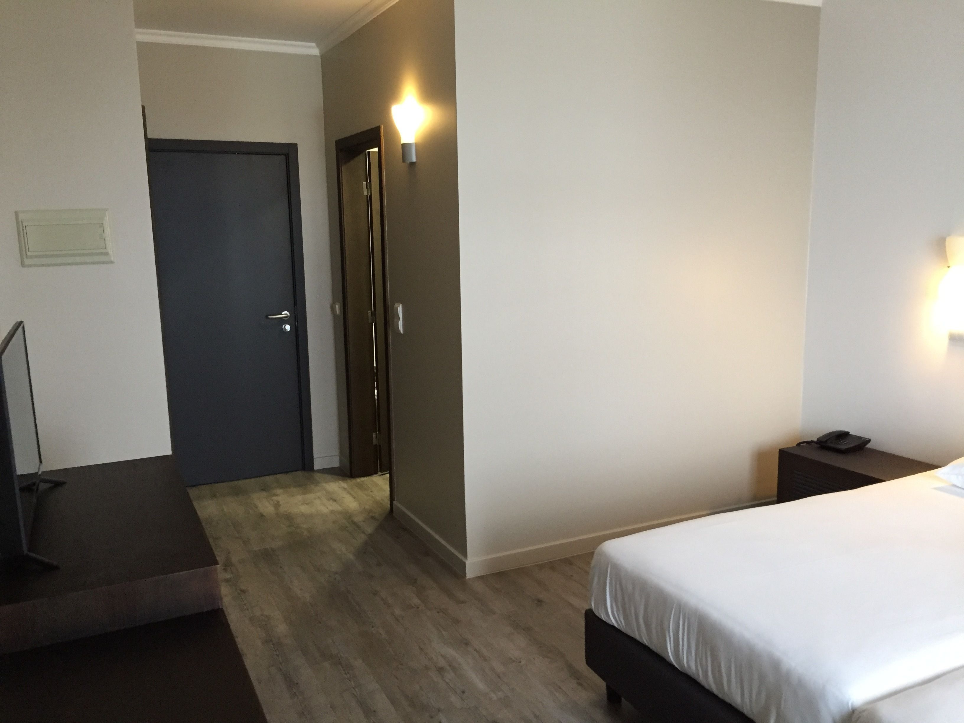 A hotel room with a bed, a desk, a chair, a door, and a wall-mounted light.