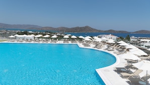 Une grande piscine claire, dotée de chaises longues et de parasols, entourée de bâtiments d’où l’on peut admirer la mer et les montagnes en arrière-plan.