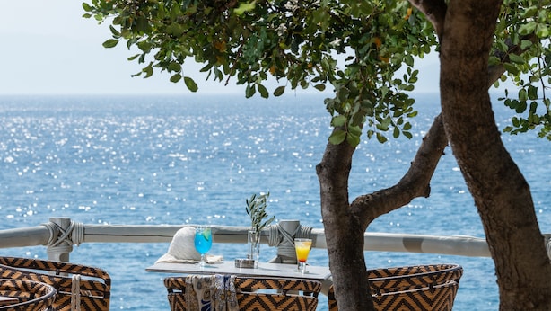 Un espace de restauration en bord de mer, avec des chaises en osier et une table dressée pour deux, offrant une vue sur l’océan.
