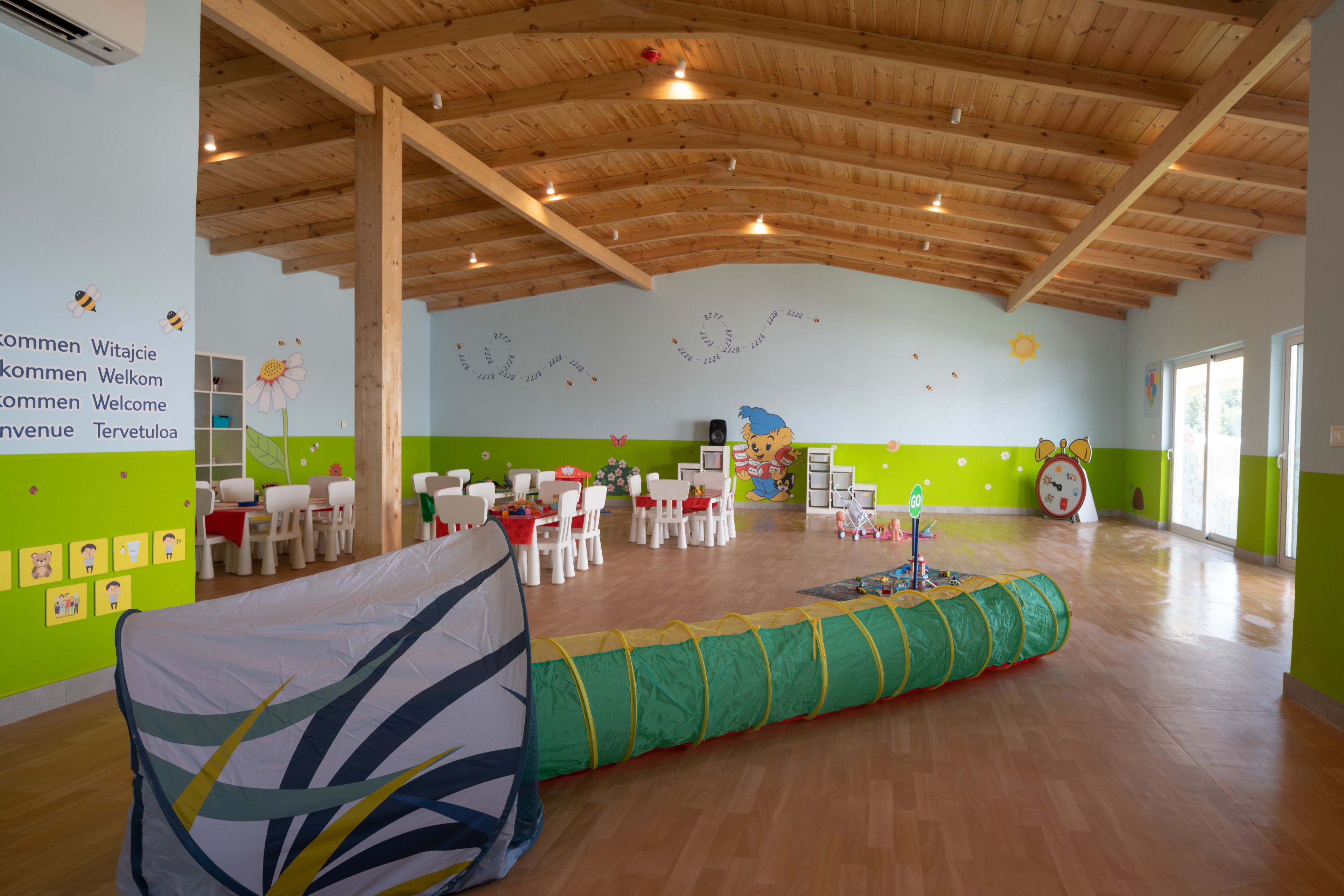 A spacious room with wooden beams on the ceiling, green walls, and a play area with a green and yellow tunnel. Tables and chairs are arranged for dining.