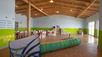 A spacious room with wooden beams on the ceiling, green walls, and a play area with a green and yellow tunnel. Tables and chairs are arranged for dining.