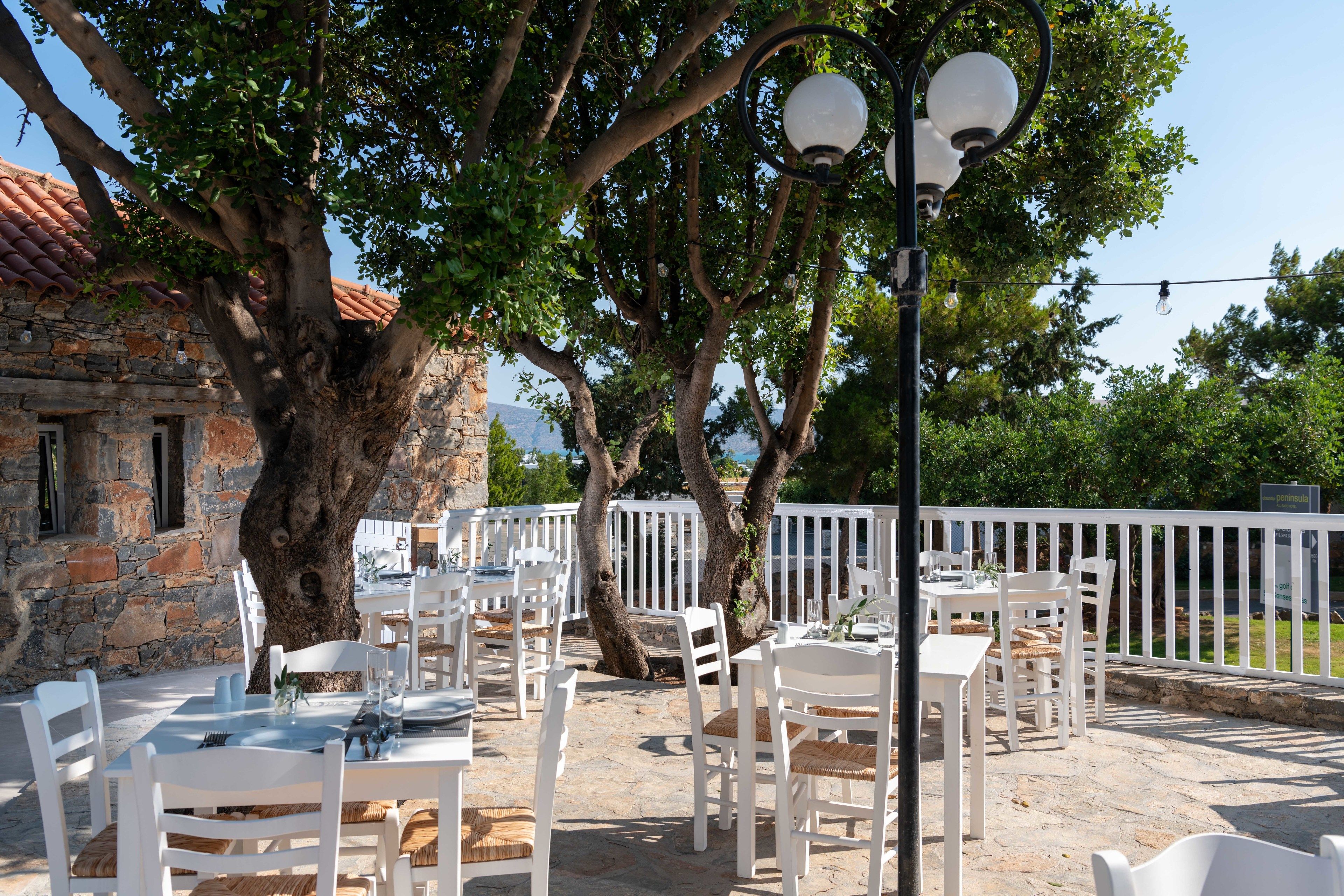 An outdoor dining area with white tables and chairs, surrounded by stone walls and trees.
