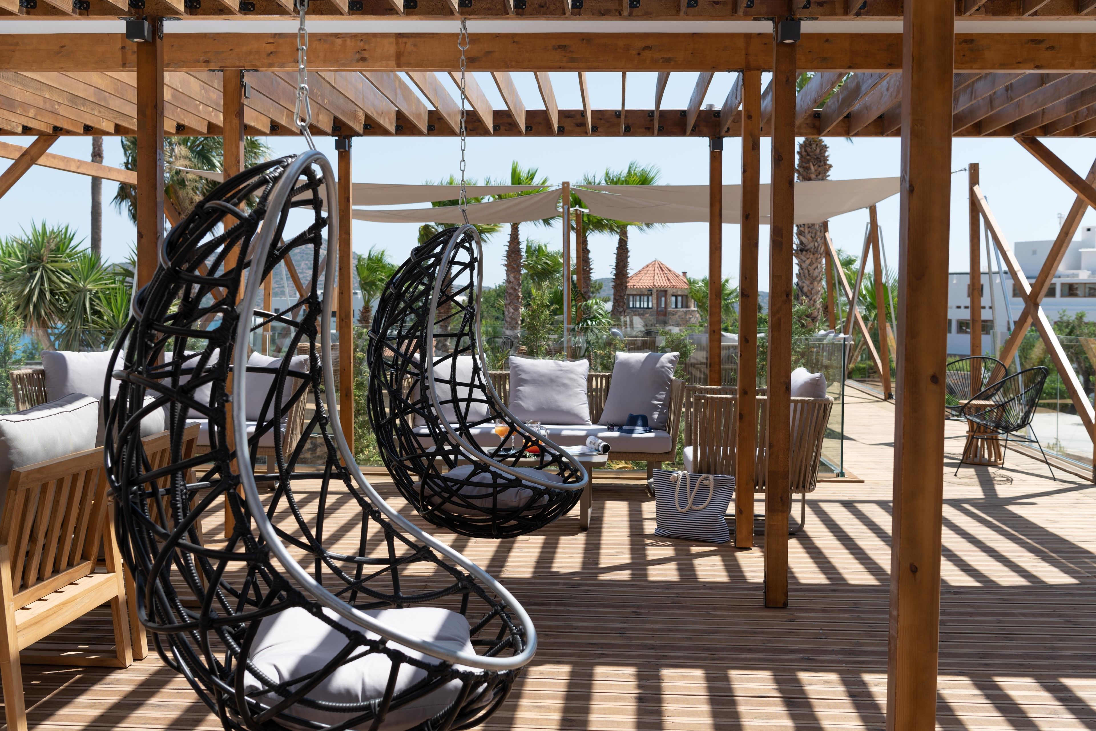 Una terraza de madera con sillas colgantes y un sofá, palmeras y un edificio al fondo.