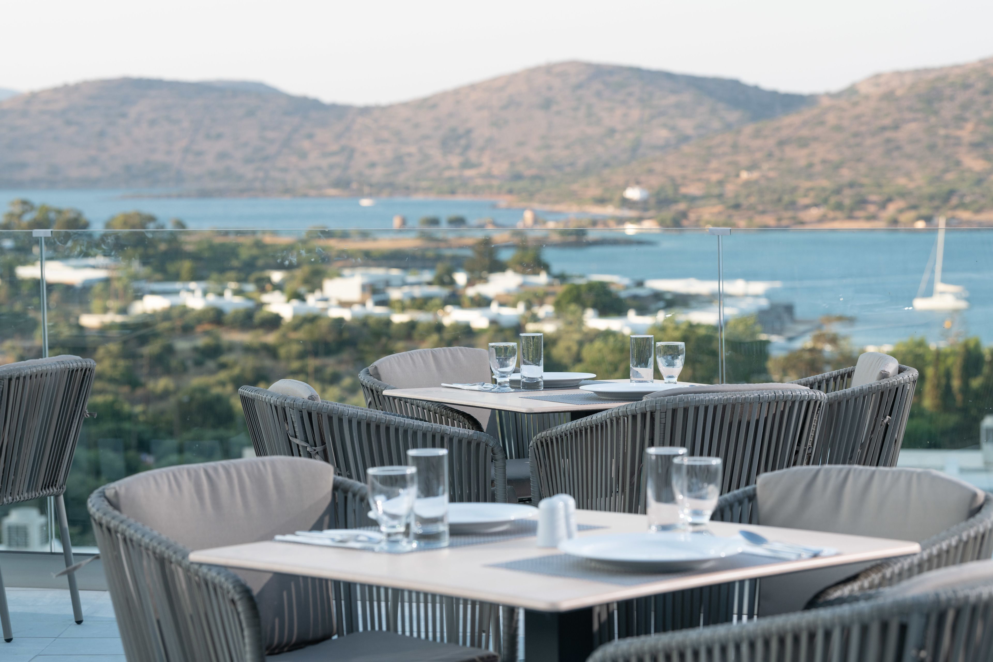 A dining area with wicker chairs and tables, overlooking a scenic view of a coastal town and hills.