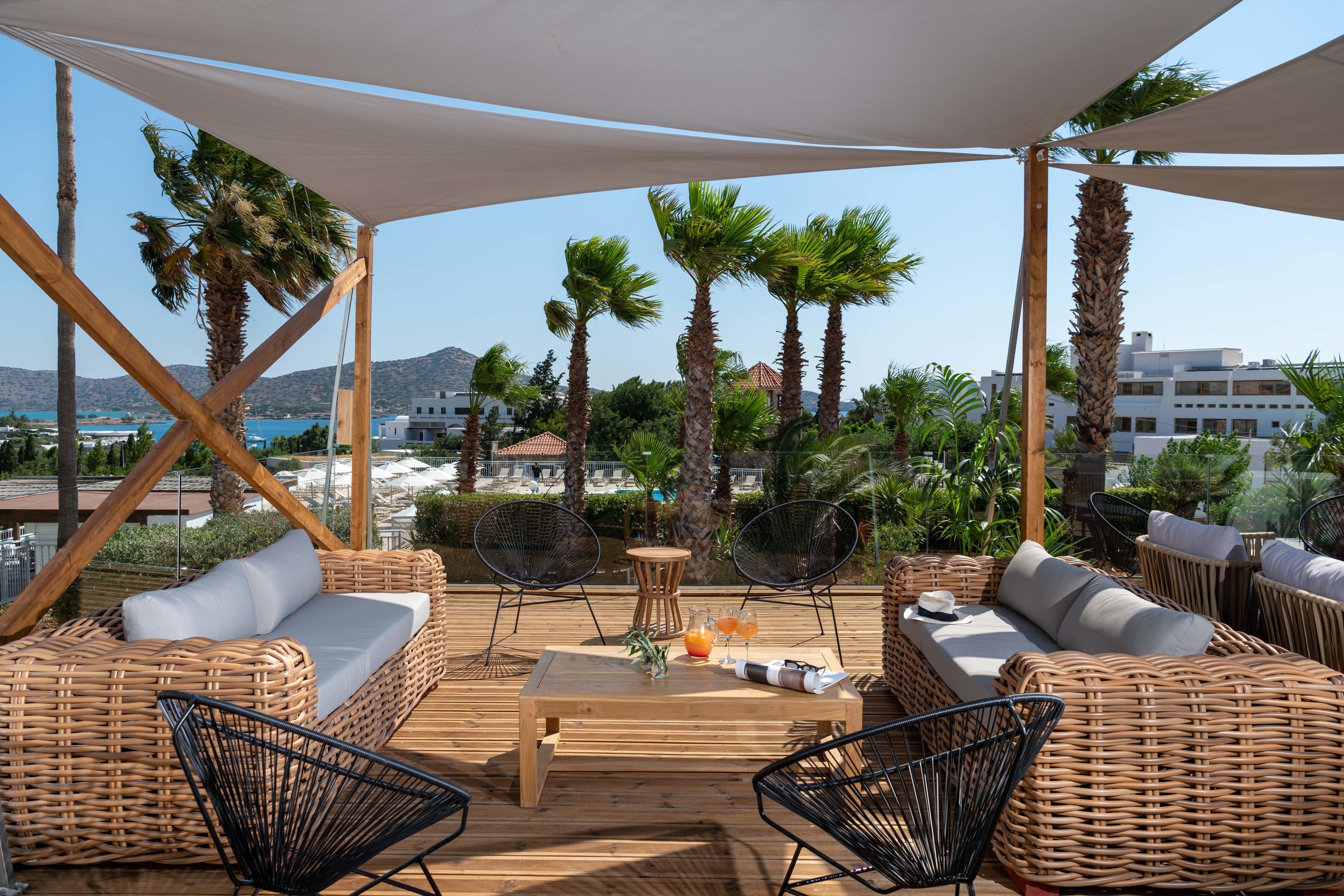 Un área de estar al aire libre con mobiliario de mimbre, una mesa de madera y vistas al mar y a edificios.