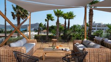 Un área de estar al aire libre con mobiliario de mimbre, una mesa de madera y vistas al mar y a edificios.