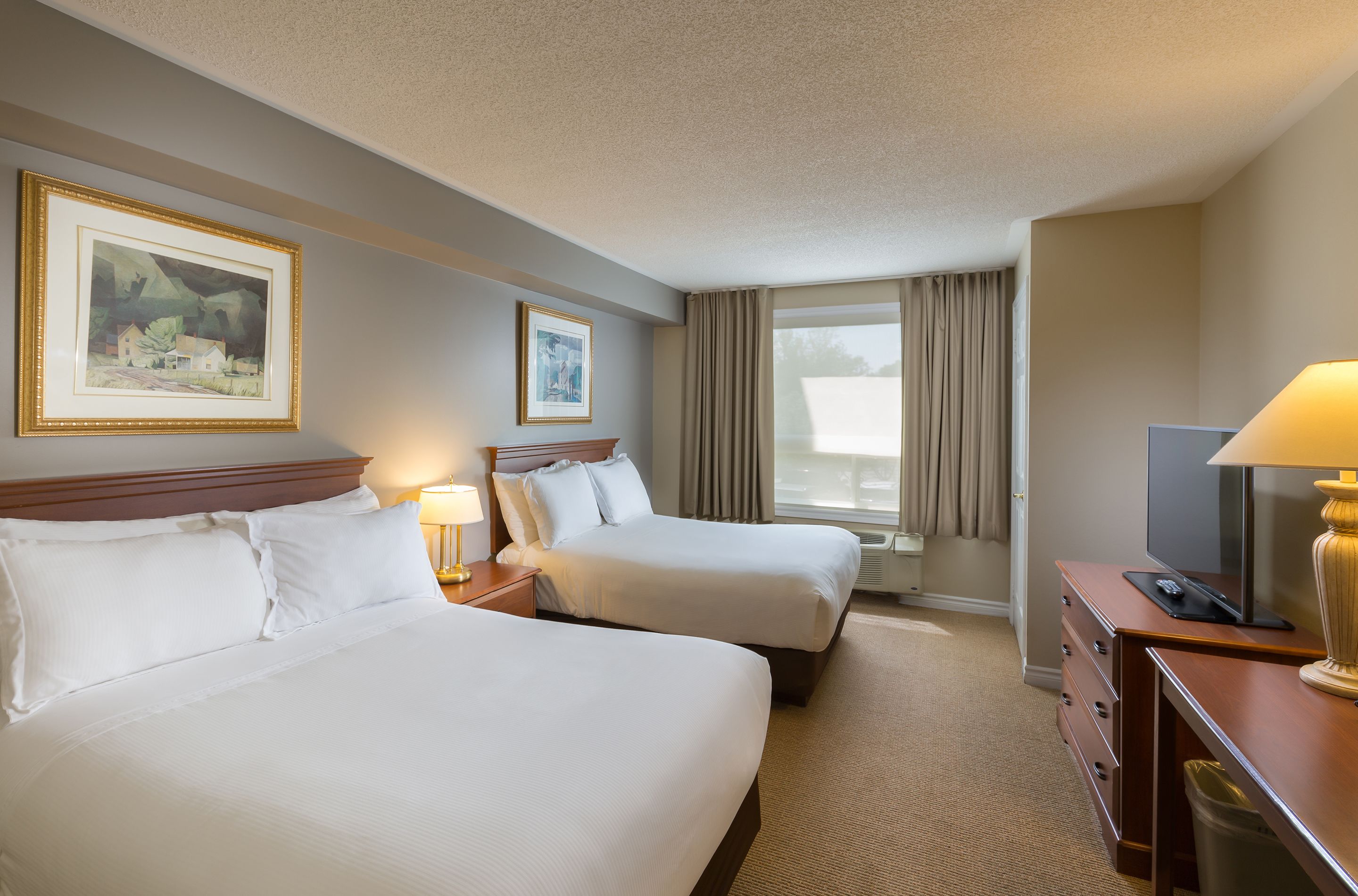 A hotel room with two beds, a desk with a lamp, a TV, and two framed pictures on the wall.