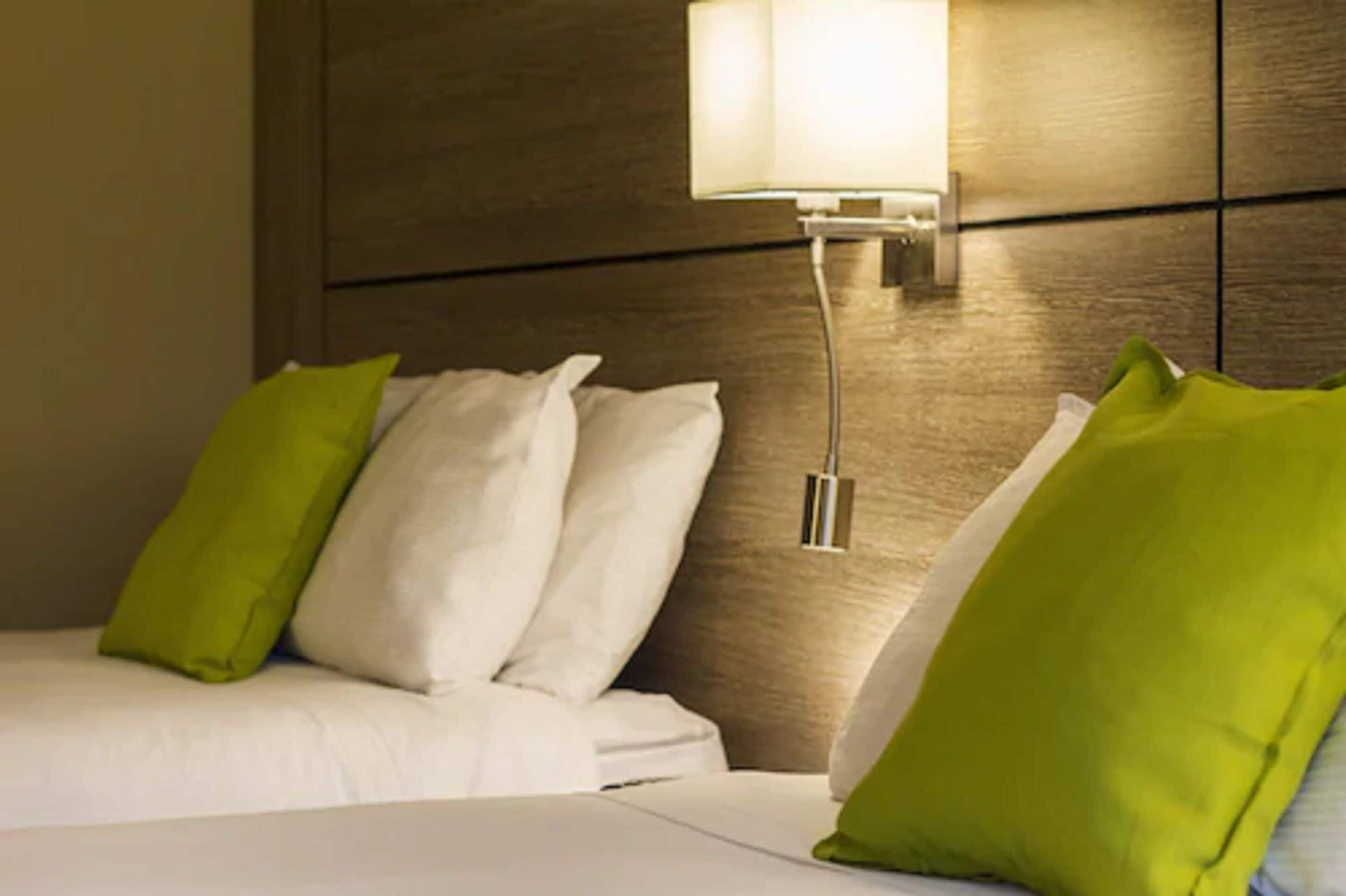 A hotel room with a bed, white bedding, and green pillows. There is a wall-mounted lamp and a wooden headboard.